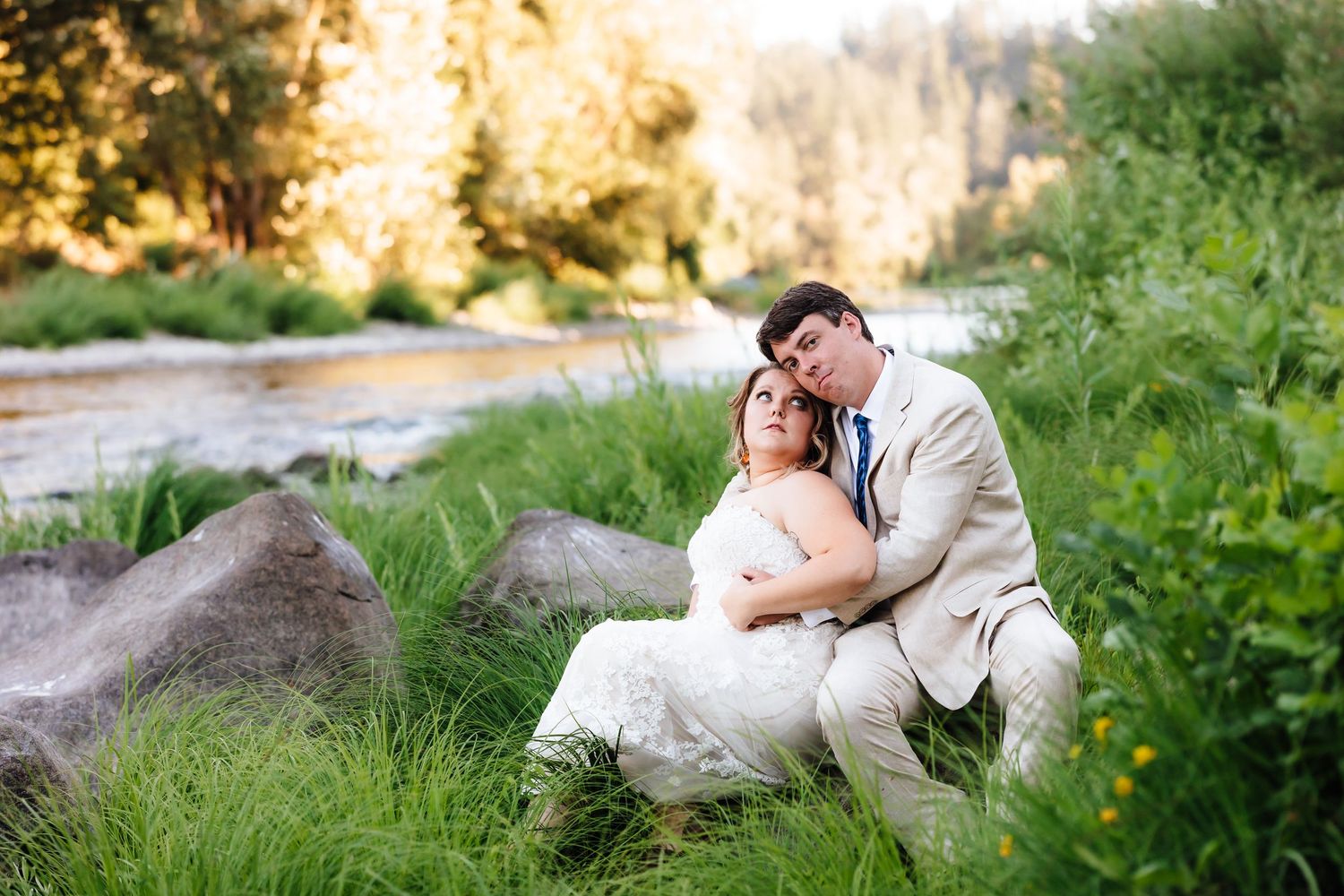 Bride and groom sharing a tender embrace beside a peaceful river surrounded by lush green grass and golden light.