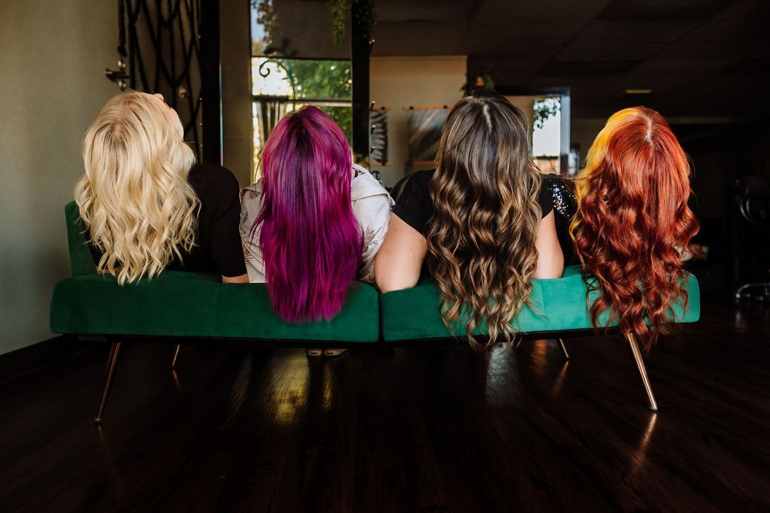 Four women with colorful hair in blonde, purple, brunette, and red sitting on green velvet sofa from behind.