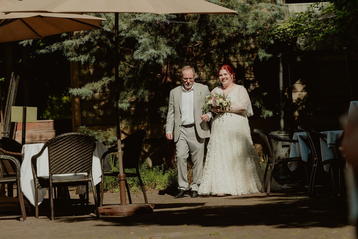 Bride in lace gown walks with older gentleman escort along outdoor wedding ceremony aisle.
