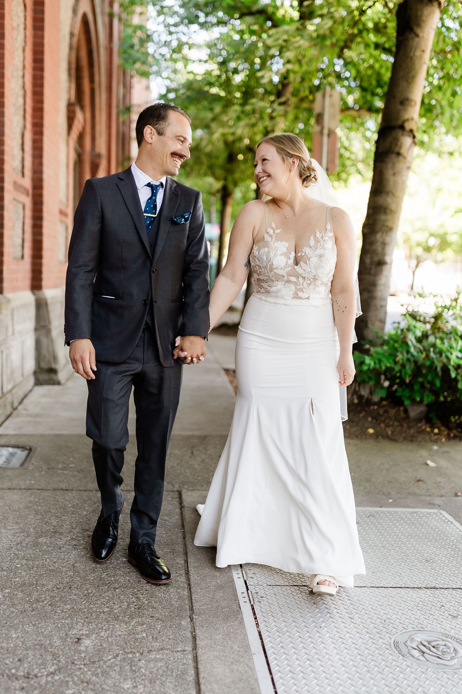 Happy bride and groom walk hand-in-hand on city sidewalk in wedding attire.