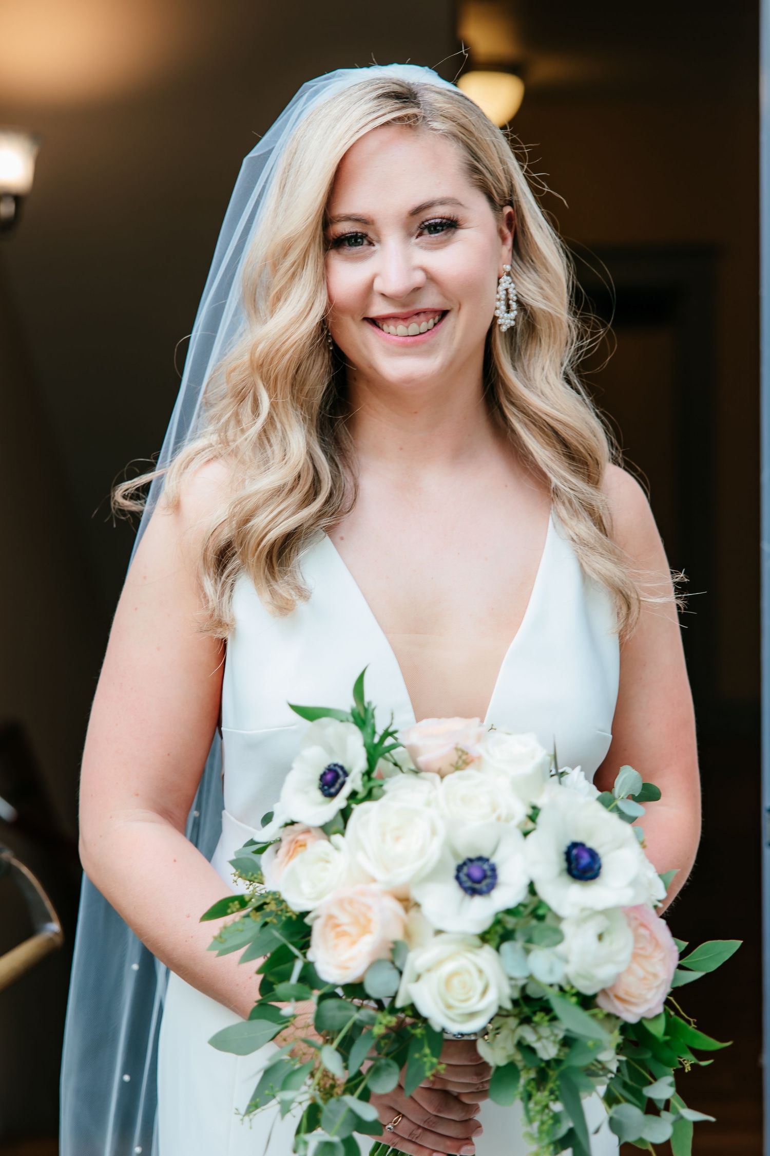 Smiling blonde bride holding a lush bouquet of white roses and blue anemones with veil.