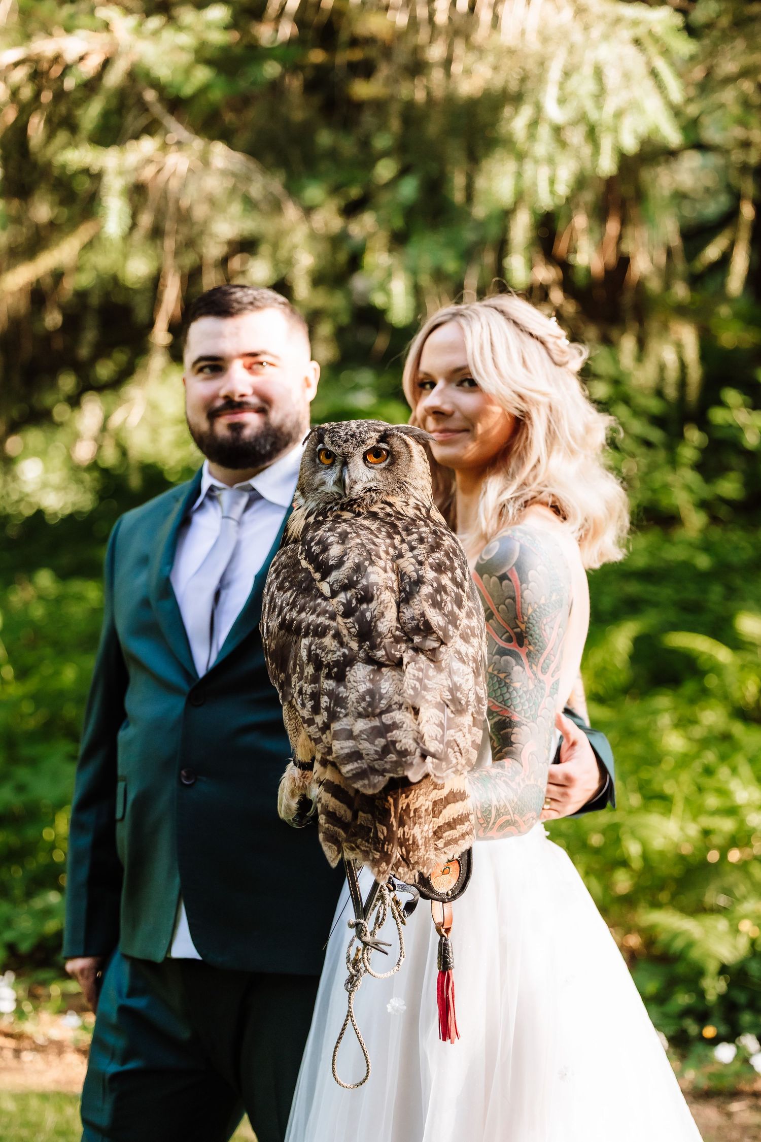 Bride with tattoo sleeve holds large owl on gloved hand beside groom in green suit outdoors.