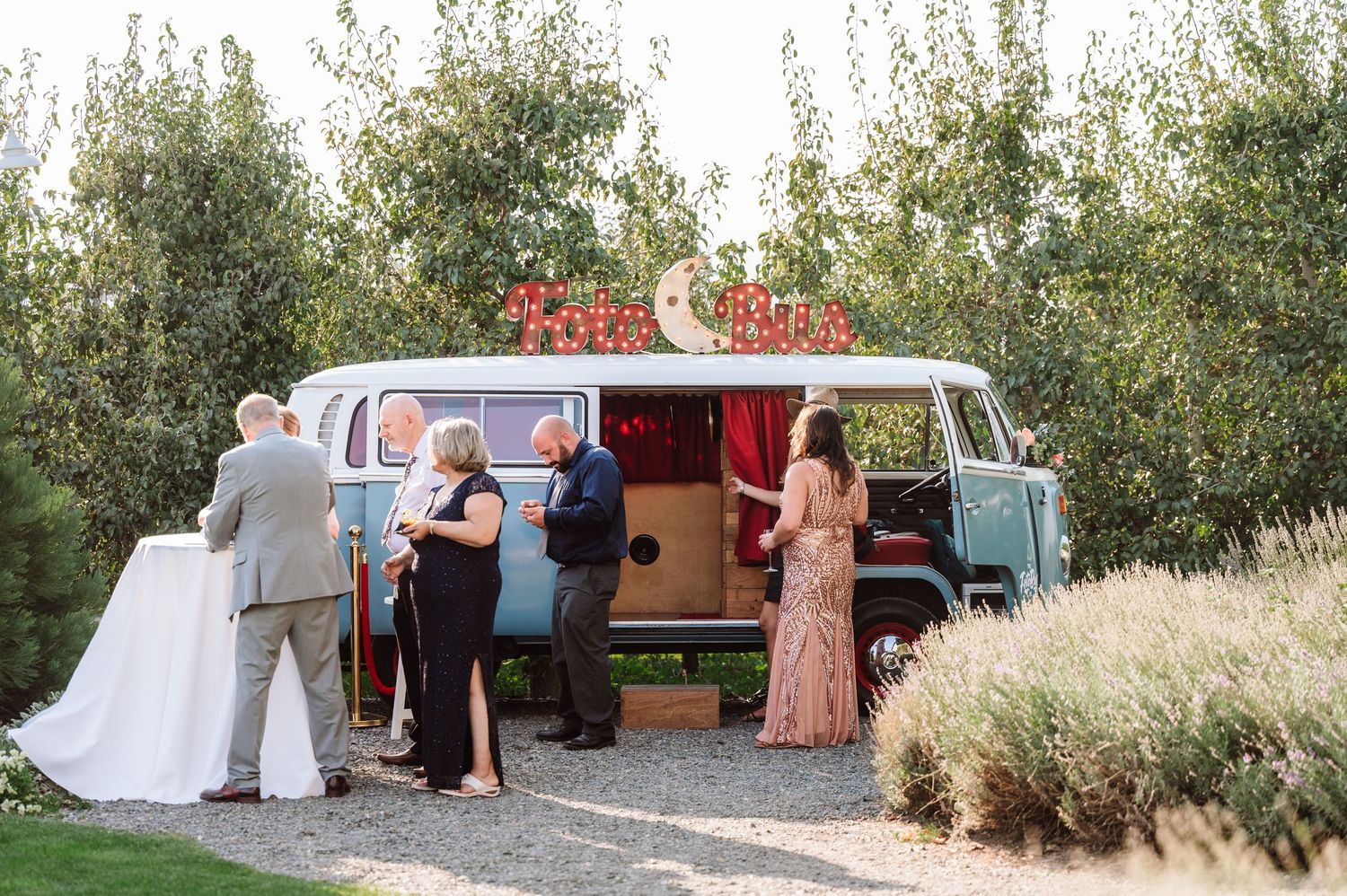 Wedding guests gather around a vintage blue VW bus photo booth at an outdoor garden reception.
