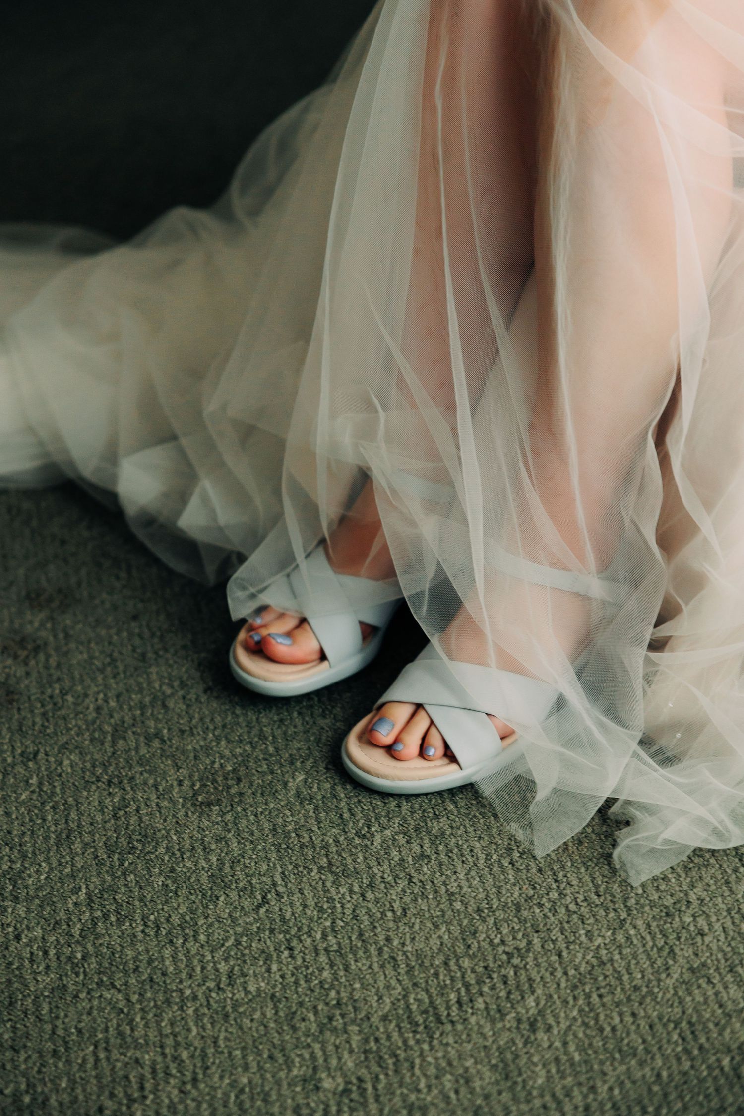 Close-up of bride's feet in white strappy sandals beneath flowing tulle wedding dress.