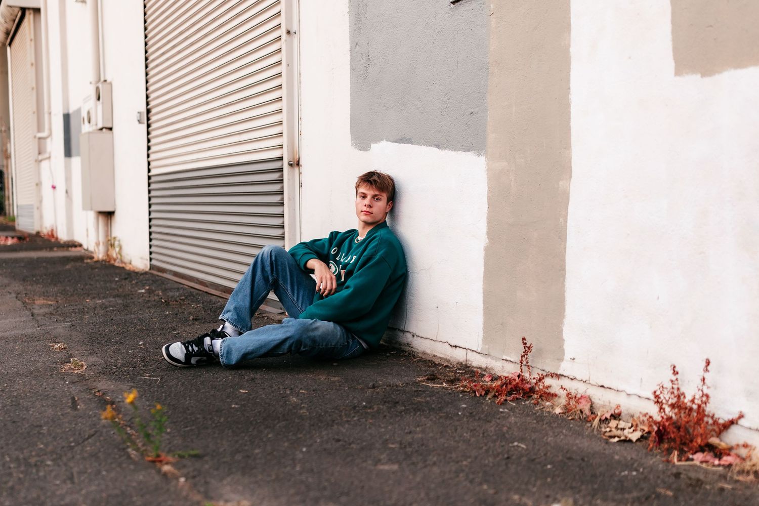 A young man in a green sweater and jeans sits casually against a white wall near a metal shutter outdoors.