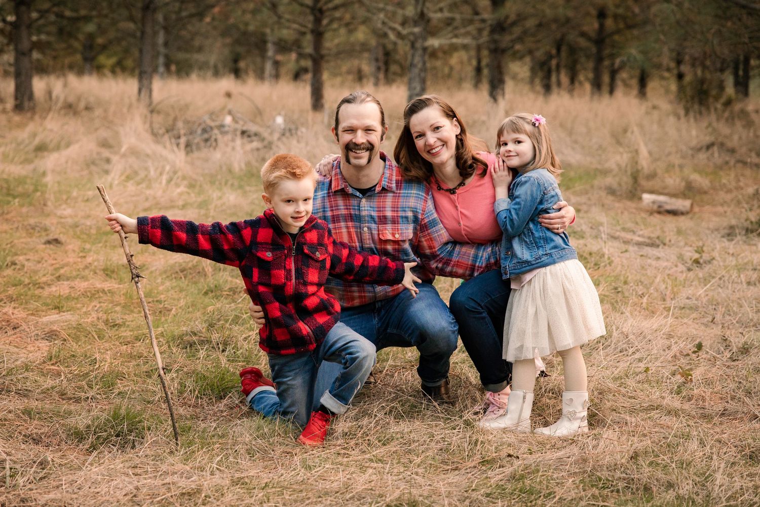 Family of four posing outdoors in a forest clearing, parents and two young children smiling in coordinating casual outfits.