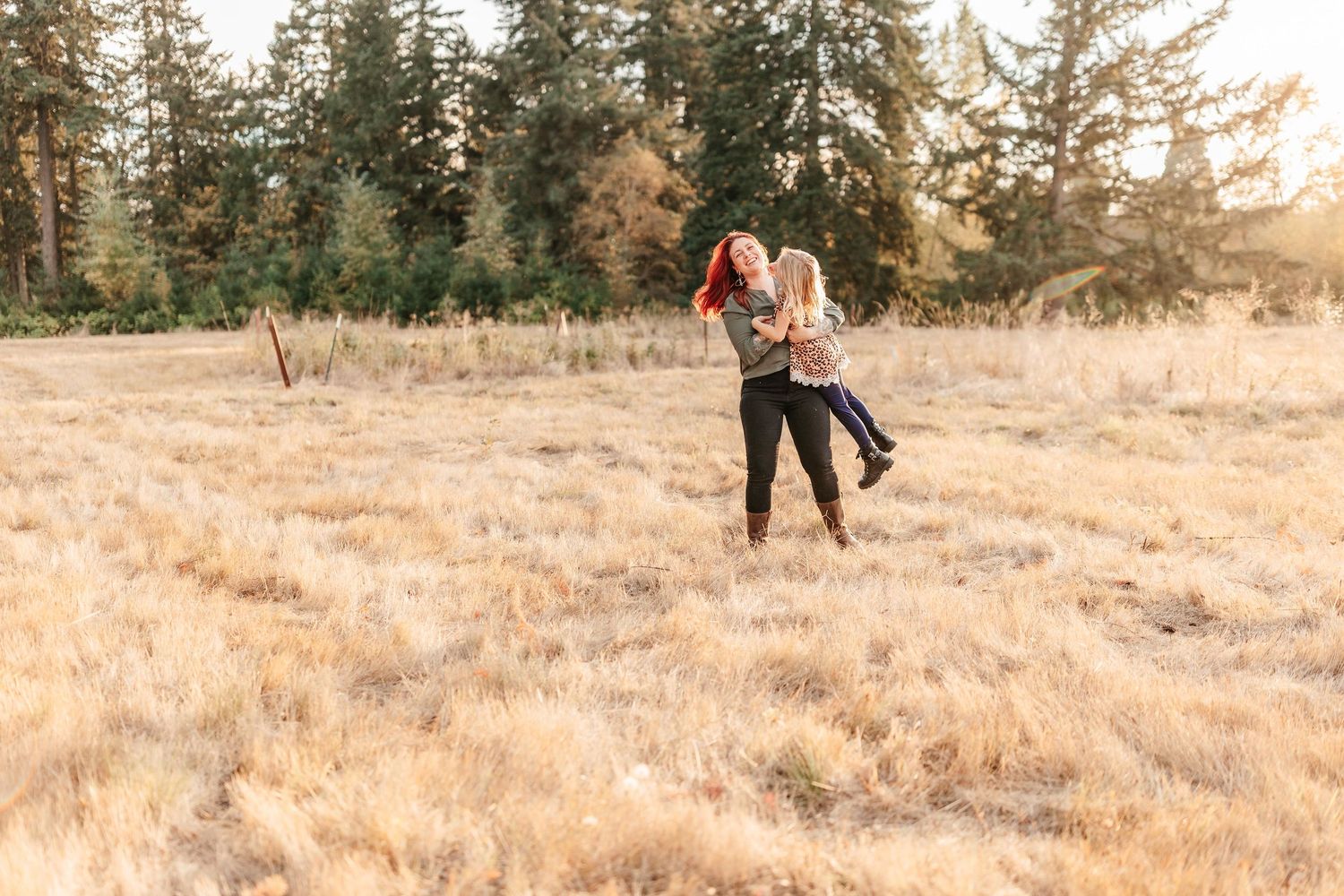 A couple embraces and spins joyfully in a golden meadow surrounded by tall pine trees during a warm sunset.