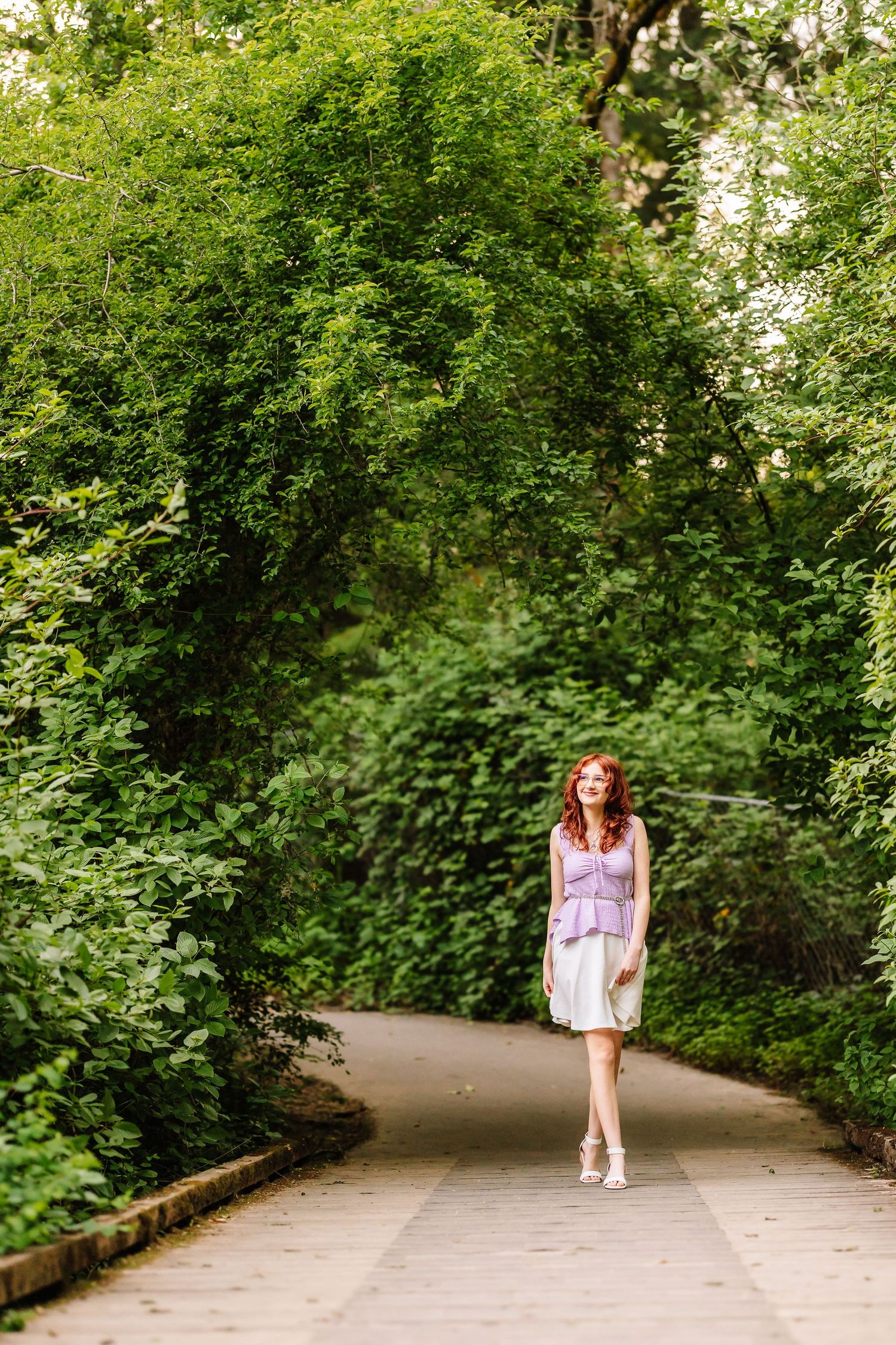 Red-haired woman in white skirt and purple top walking on a path through a lush green garden archway.