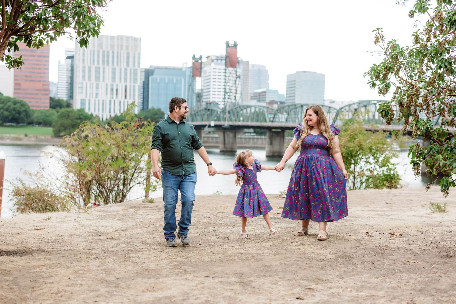Family of three holding hands and walking near a river with a city skyline and bridge in the background.