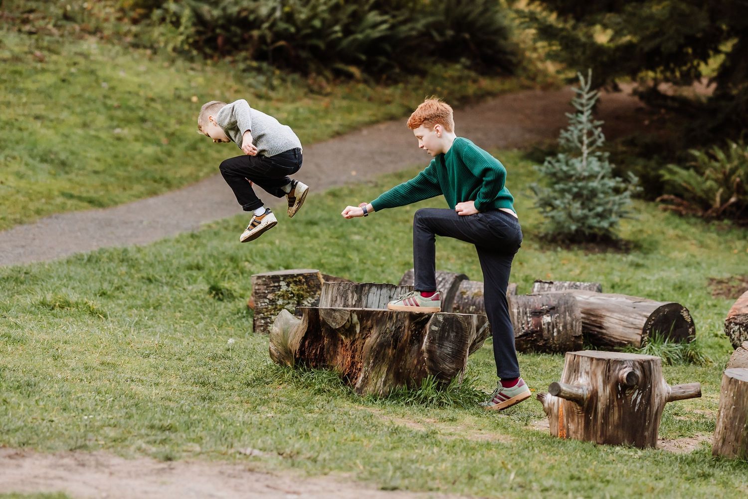 Two children playing on large logs in a green park, one jumping mid-air while the other climbs over the wooden obstacles.