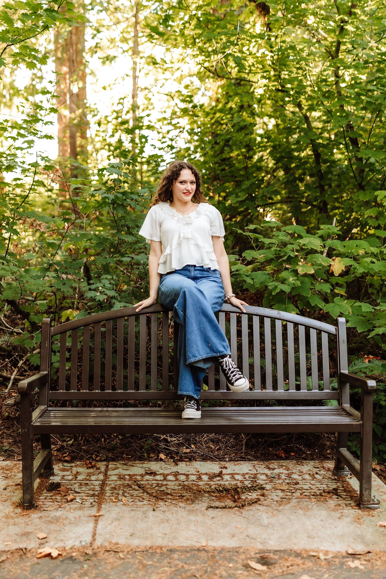 Young woman in white top and jeans sitting on a wooden park bench surrounded by lush green trees.