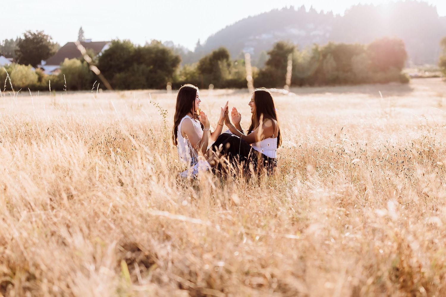 Two young women sitting face to face in golden wheat field with sunlit hills in background.