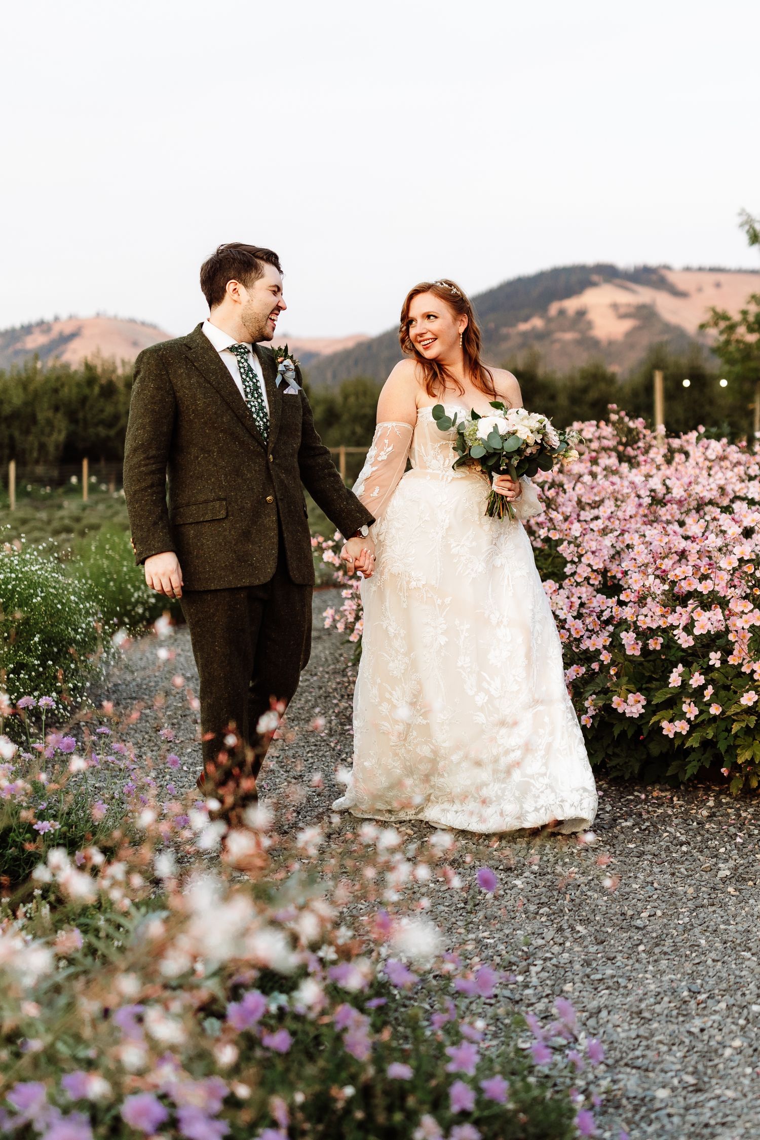 Smiling newlyweds walk hand in hand through colorful flower garden with mountain backdrop.