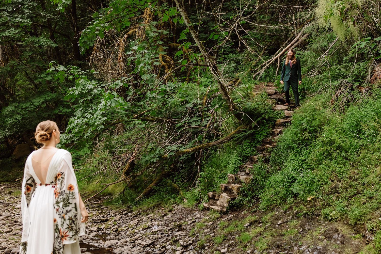 Bride in floral gown walks rocky path as groom descends stone steps in a lush forest setting.