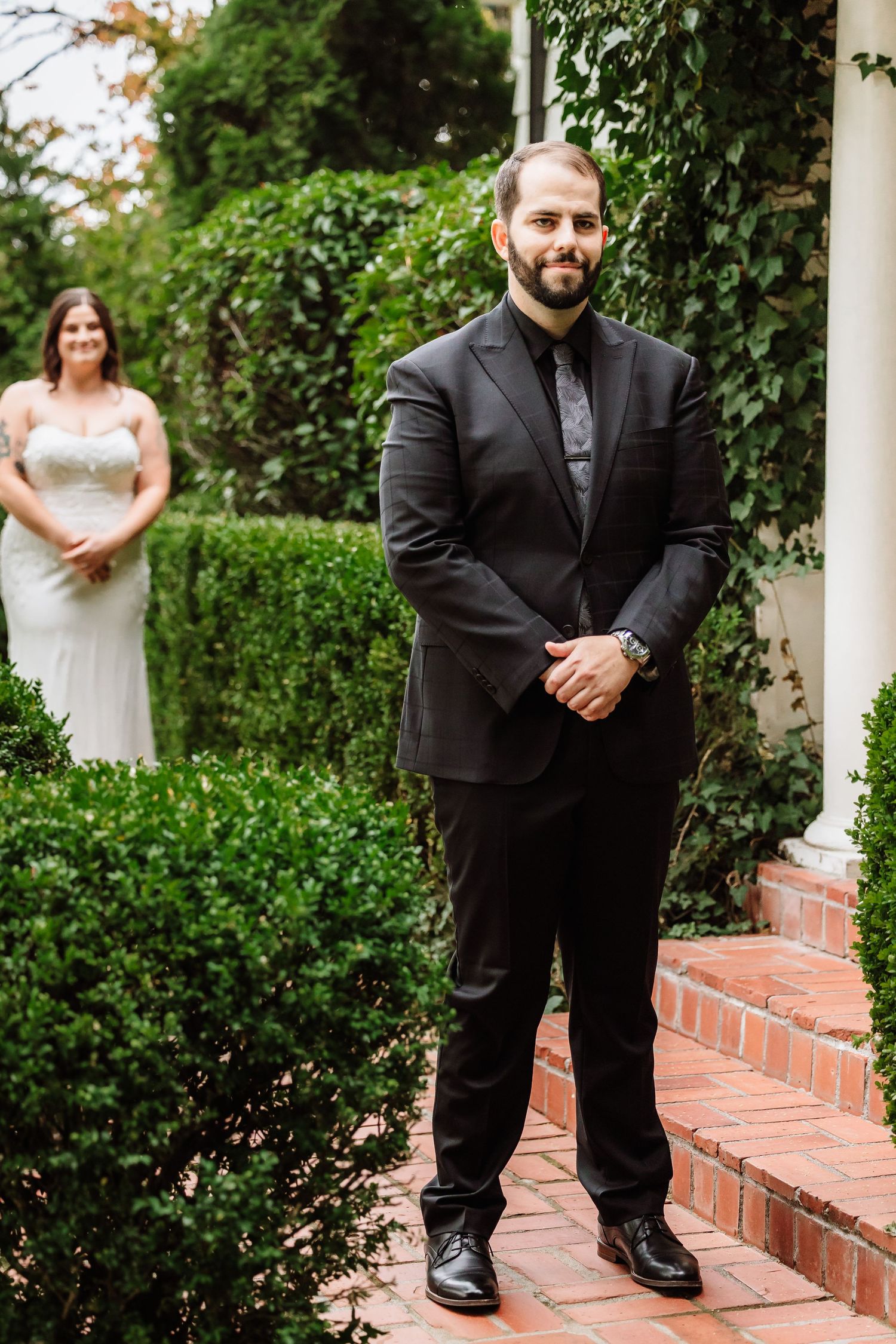 Groom in black suit smiling with bride standing in lush green garden background.