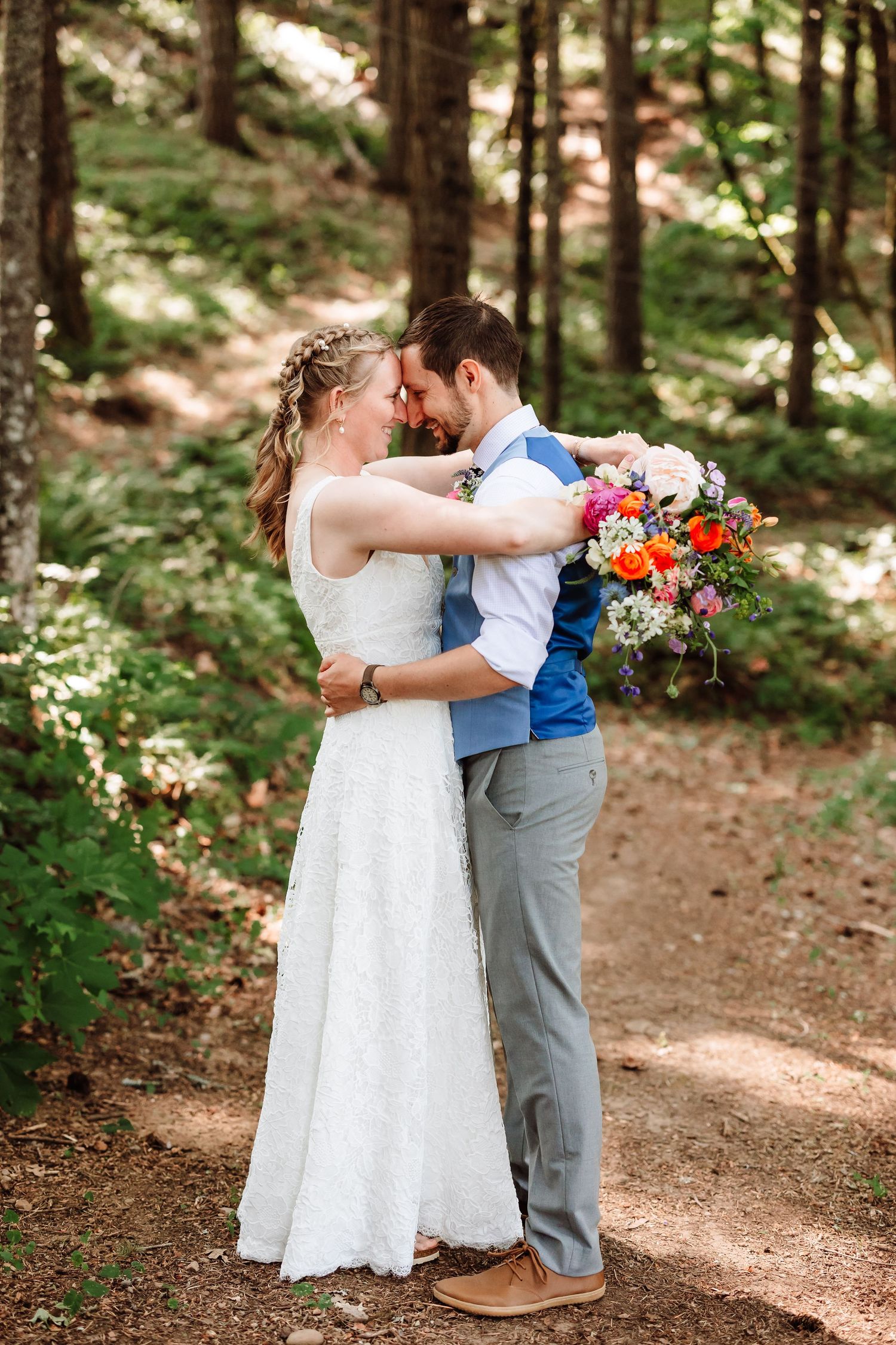 Bride and groom touch foreheads tenderly in a sunlit forest, bride holding colorful wildflower bouquet.