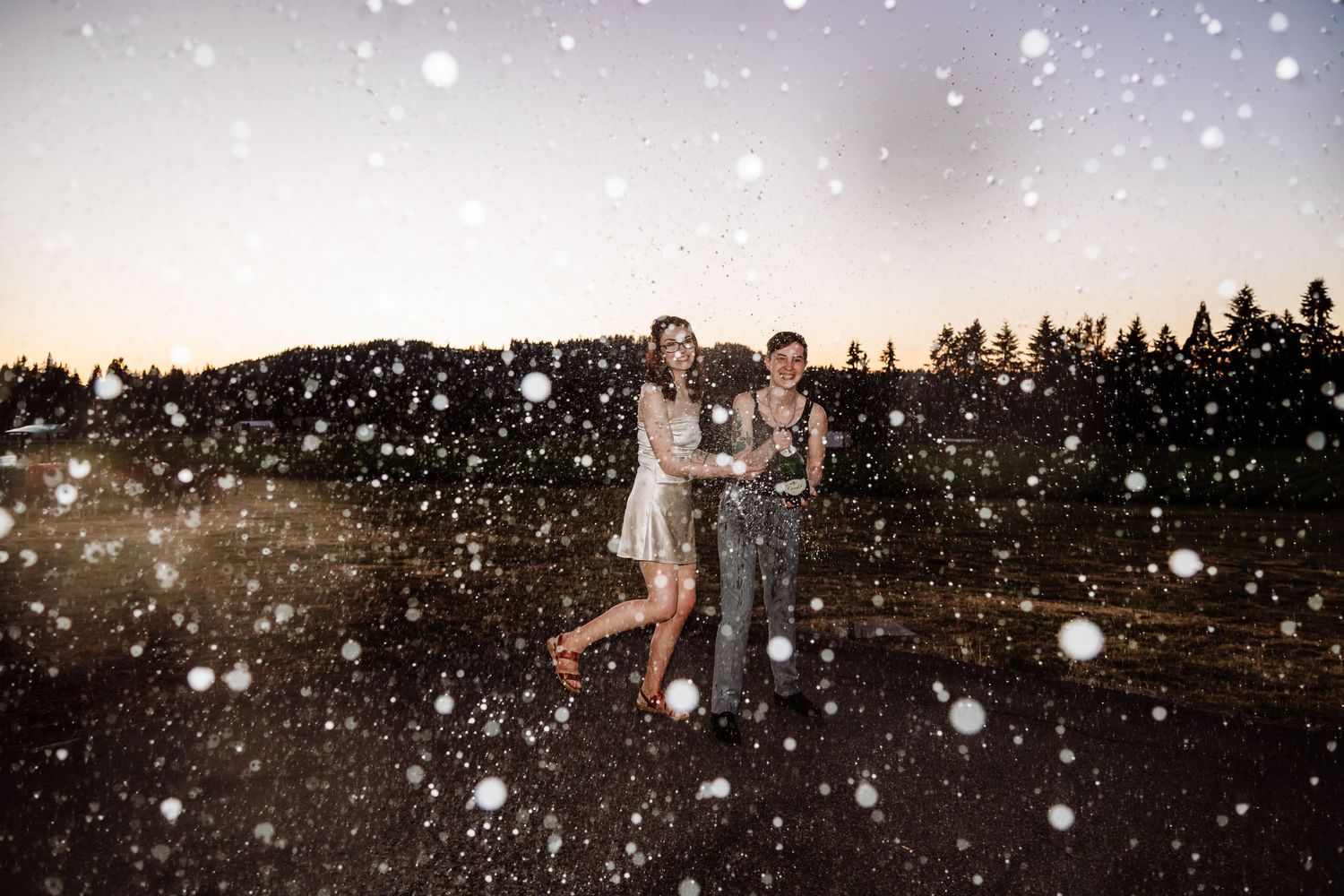 Two people laughing and dancing in snow flurries at twilight with forest backdrop.