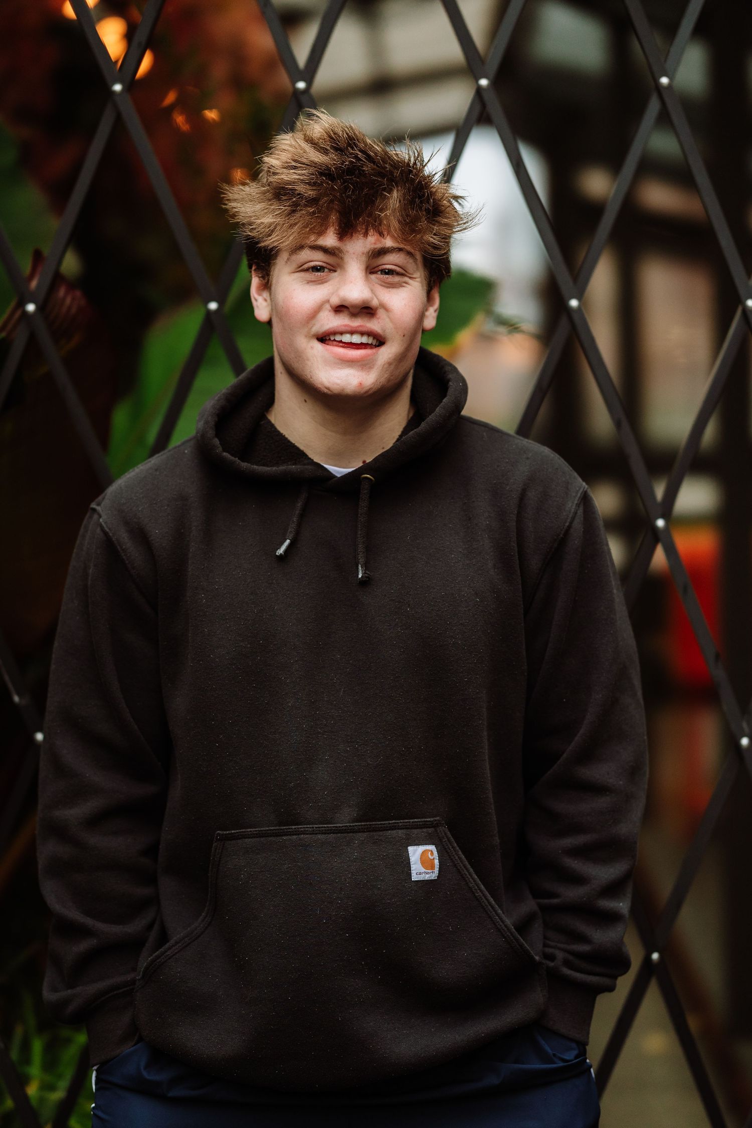 Smiling teenage boy with tousled hair wearing a dark Carhartt hoodie, standing outdoors in front of a metal lattice fence.