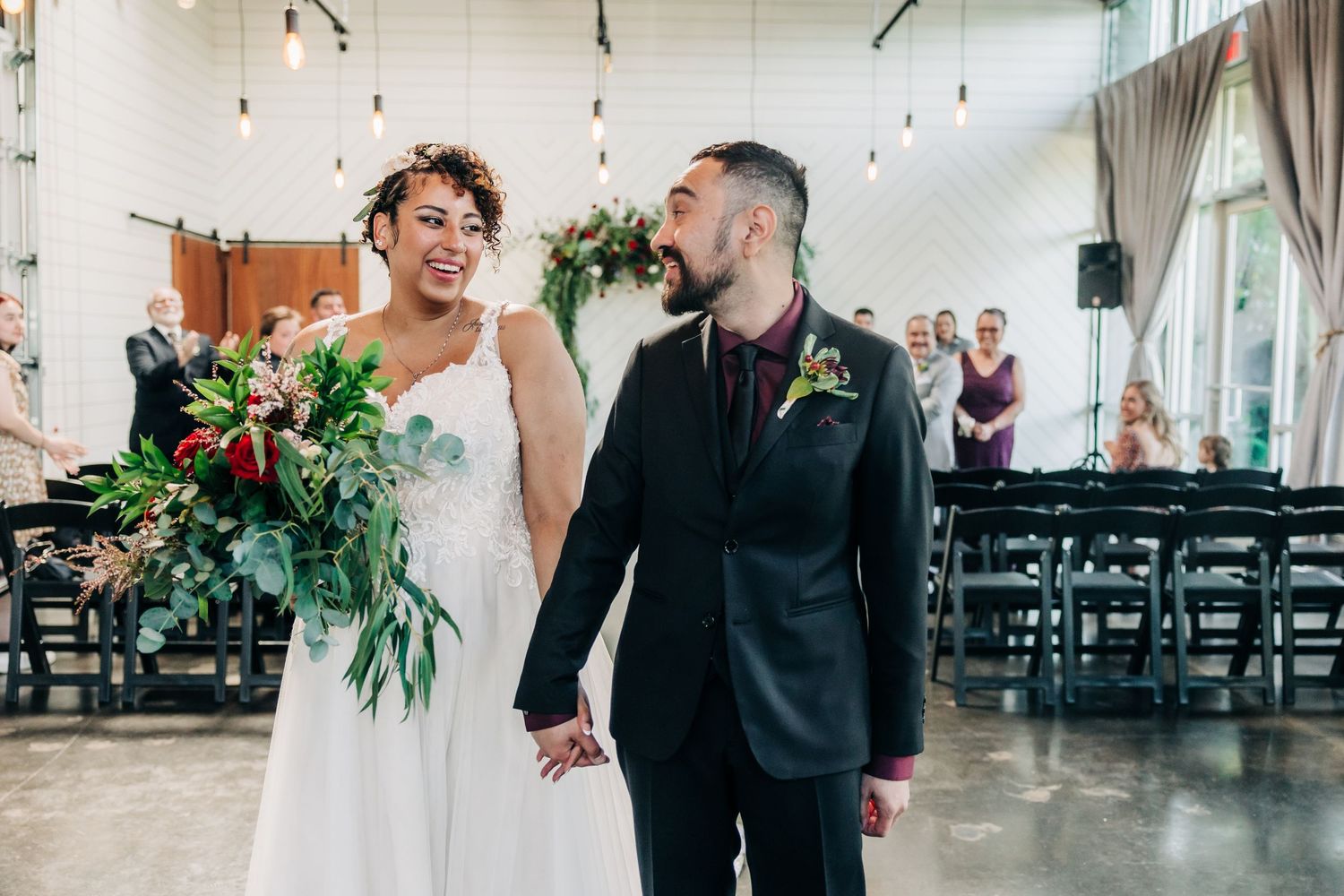 Newlyweds hold hands and smile walking back down aisle after indoor wedding ceremony.