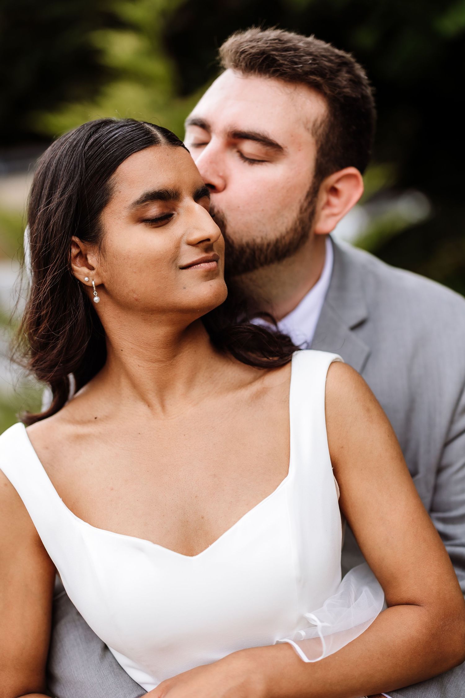 Groom tenderly kissing bride on the cheek as she smiles with eyes closed outdoors.