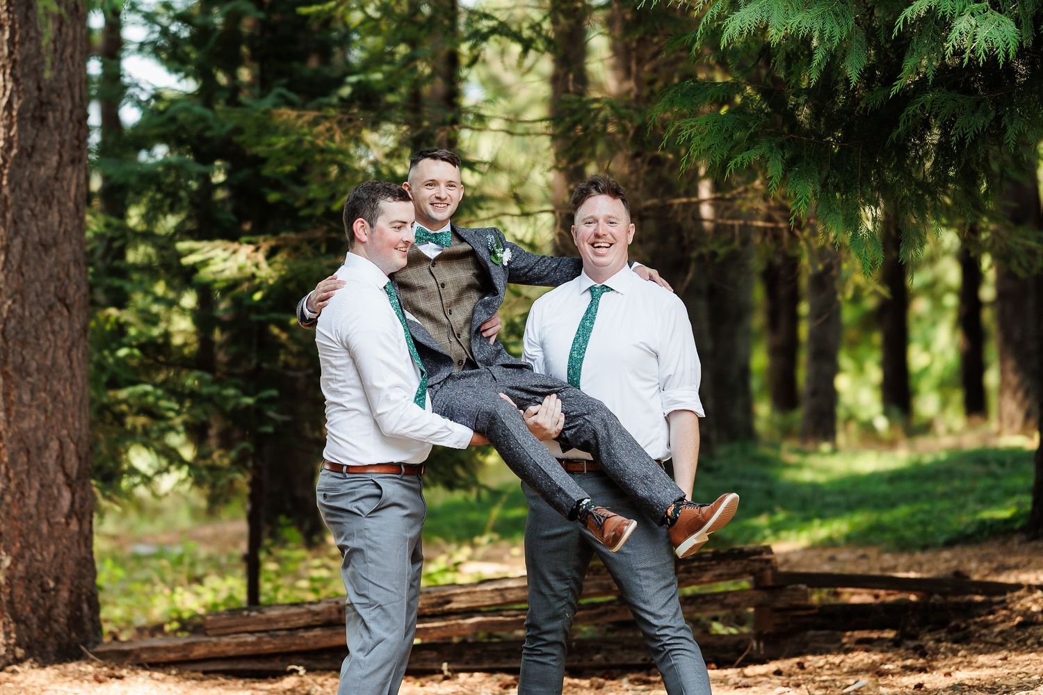 Two groomsmen playfully lift groom in forest setting, all wearing white shirts with green and teal neckties.