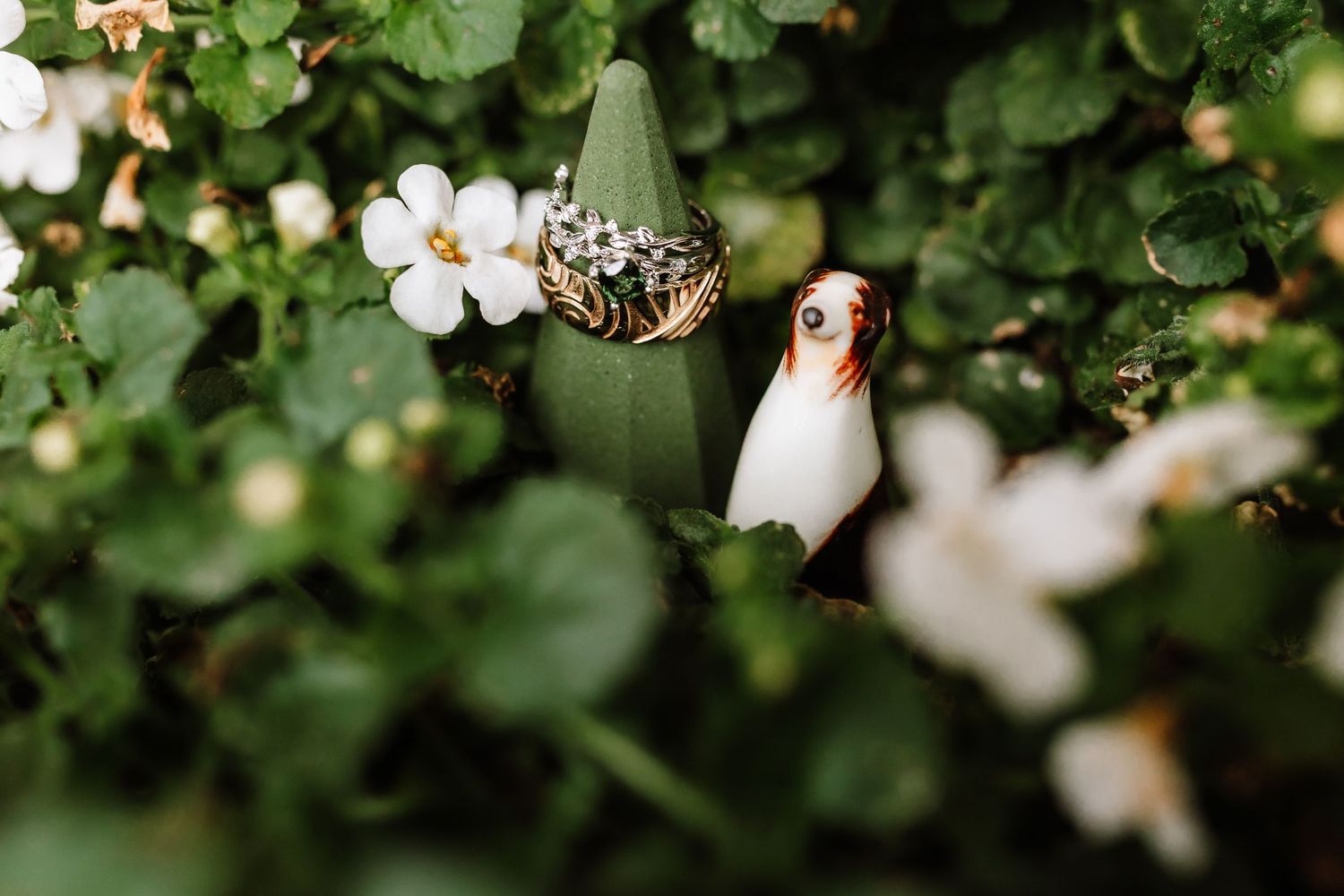 Delicate wedding ring and ceramic bird figurine nestled among white flowers and green foliage.
