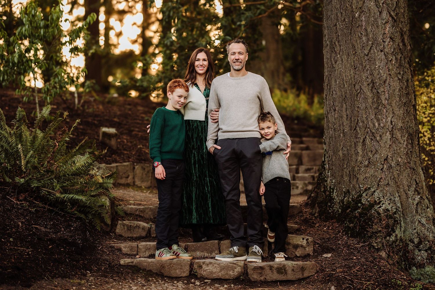 Family of four smiling outdoors on stone steps surrounded by trees in a warm autumn forest setting.