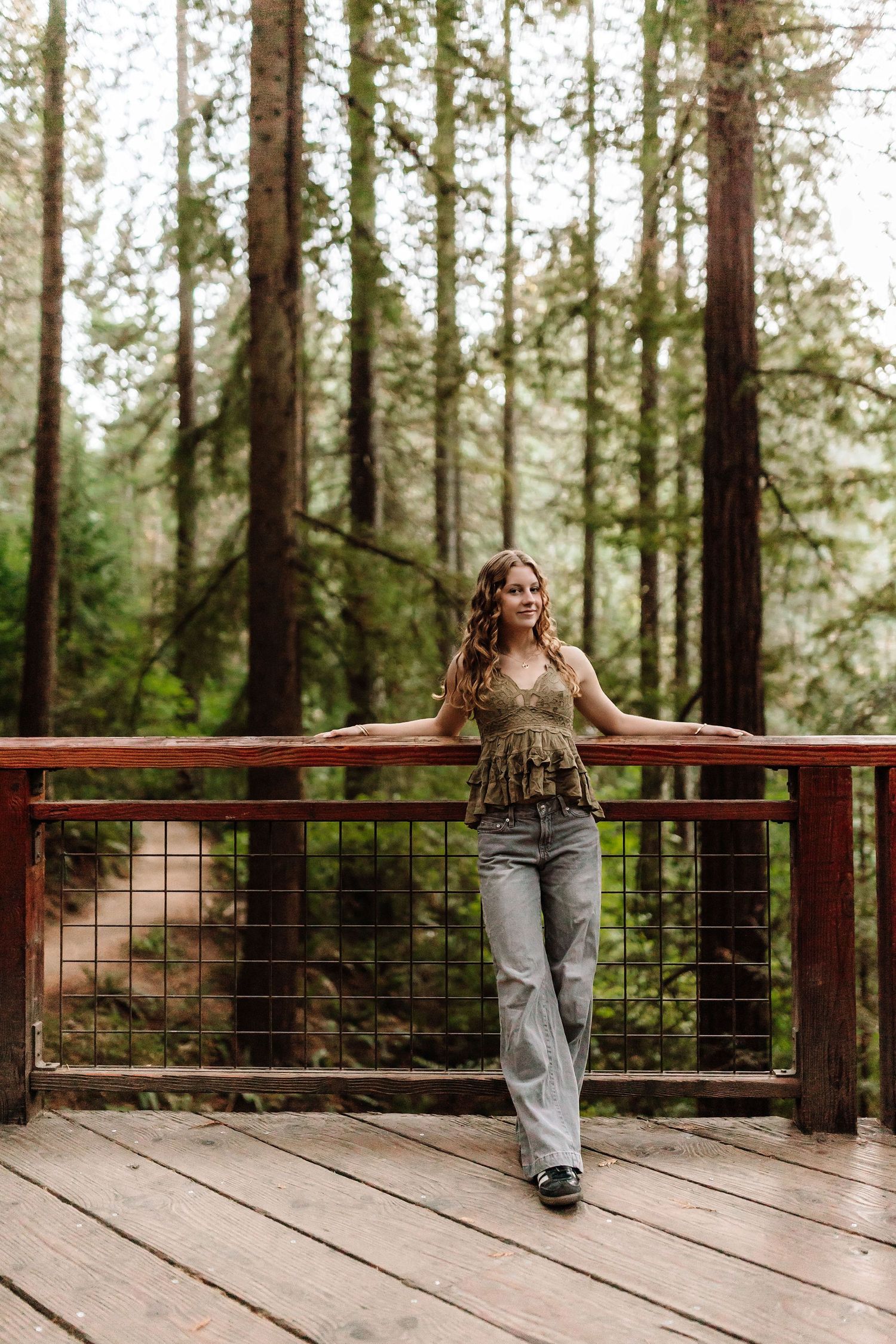 Woman in olive top and jeans leaning on wooden railing of forest deck surrounded by tall redwood trees.