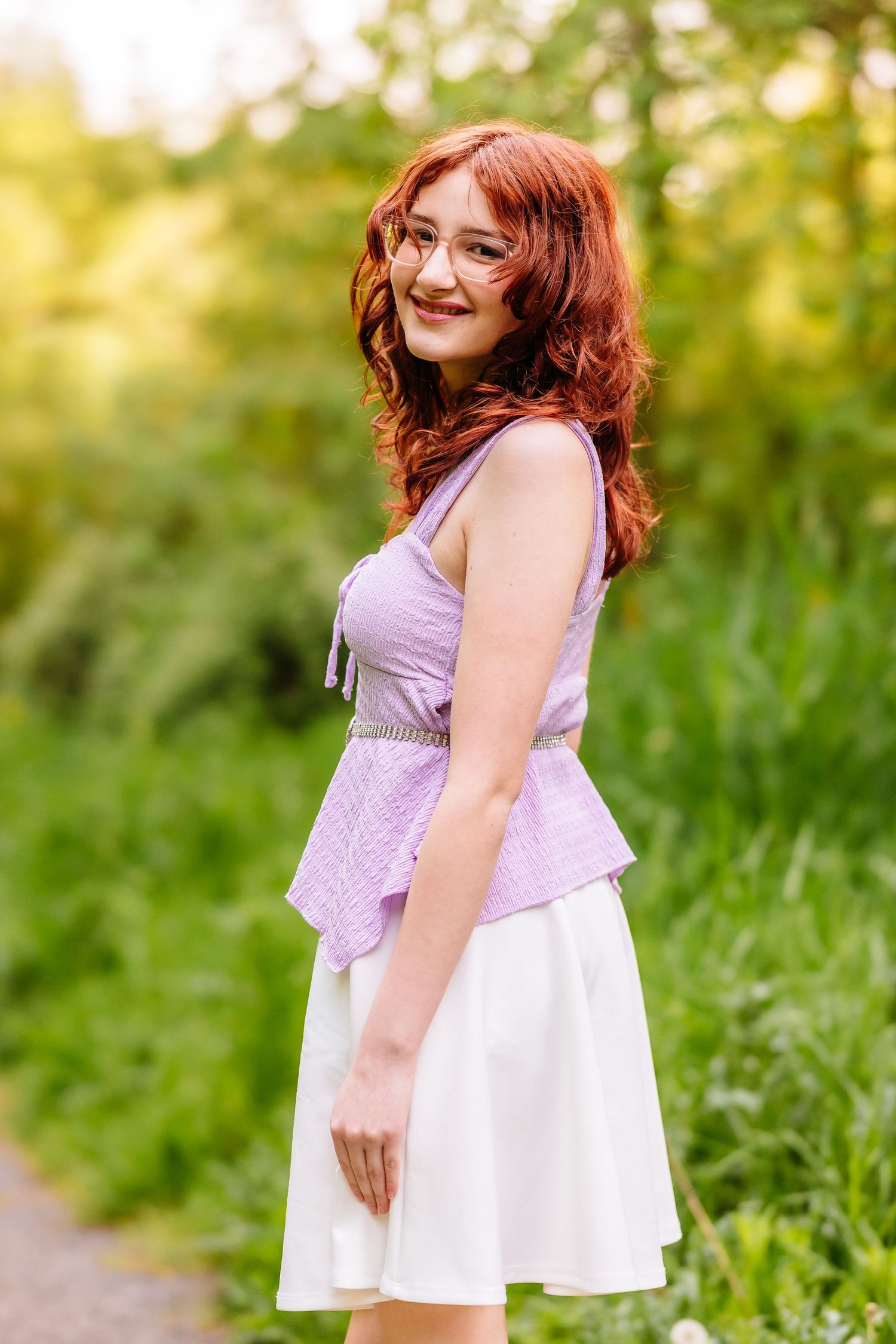 Young redhead woman in purple top and white skirt smiling outdoors in a lush green park setting.