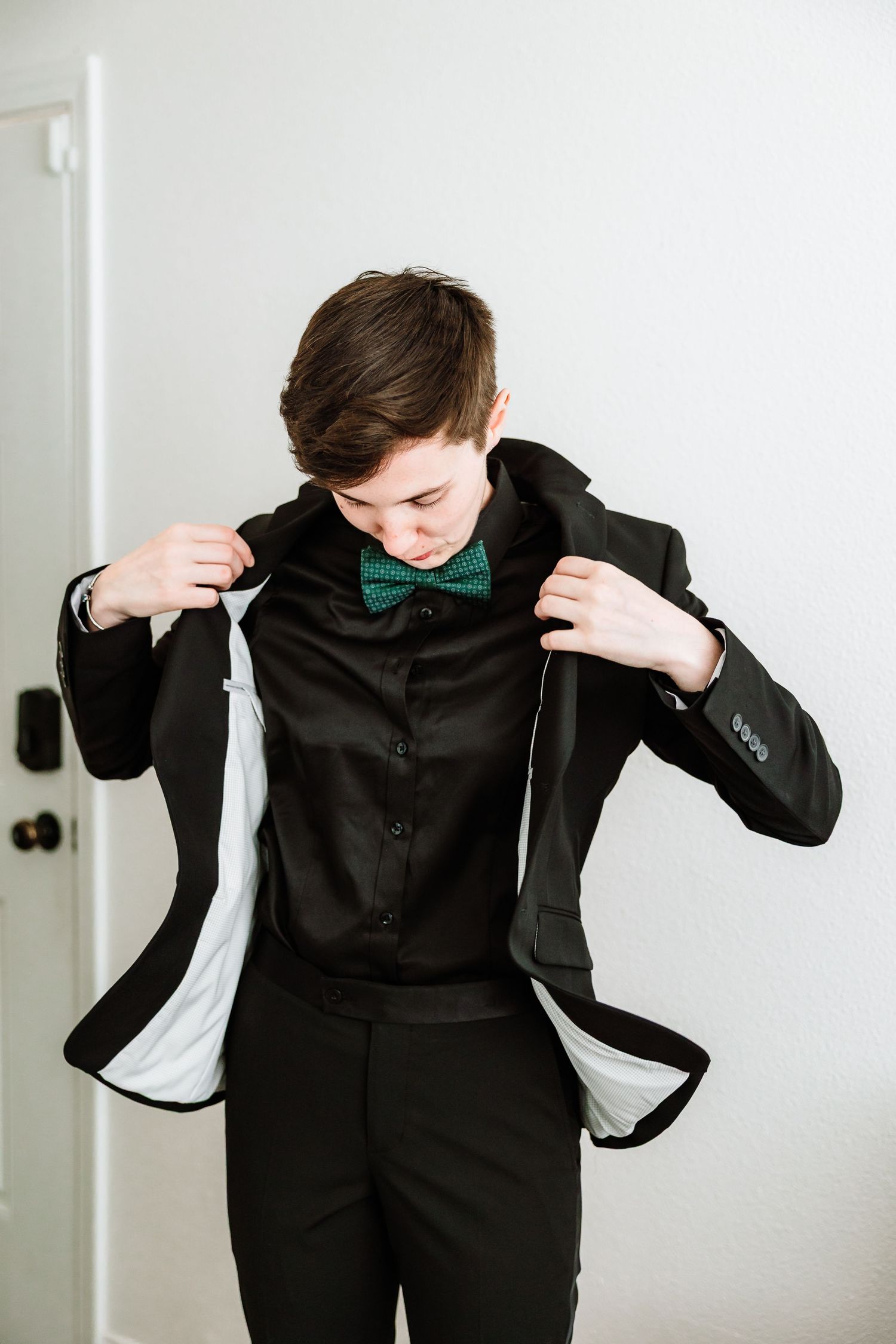 Young groom in black tuxedo with green bow tie adjusts his jacket before the wedding.