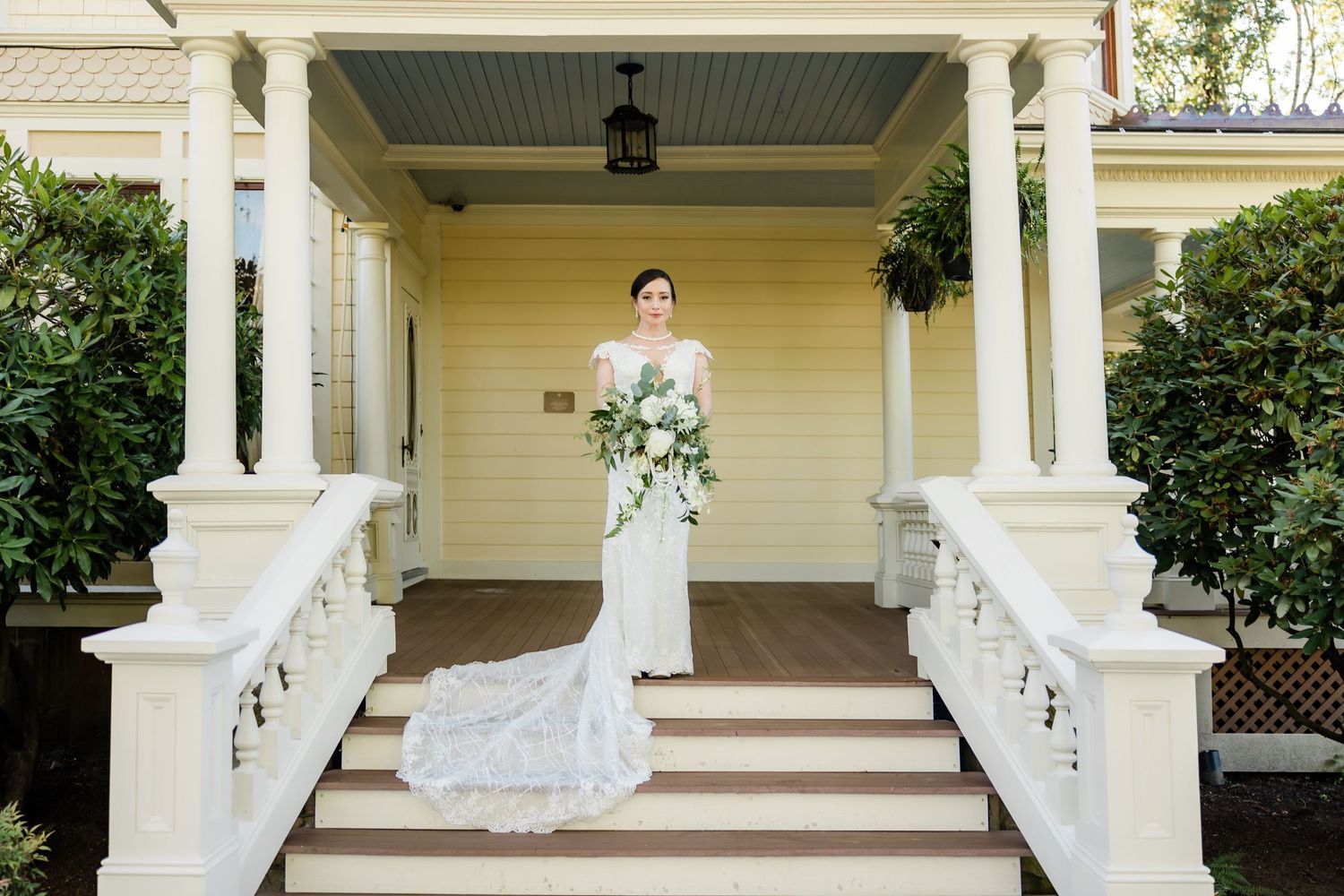Elegant bride in lace gown holds bouquet on steps of yellow Victorian home.