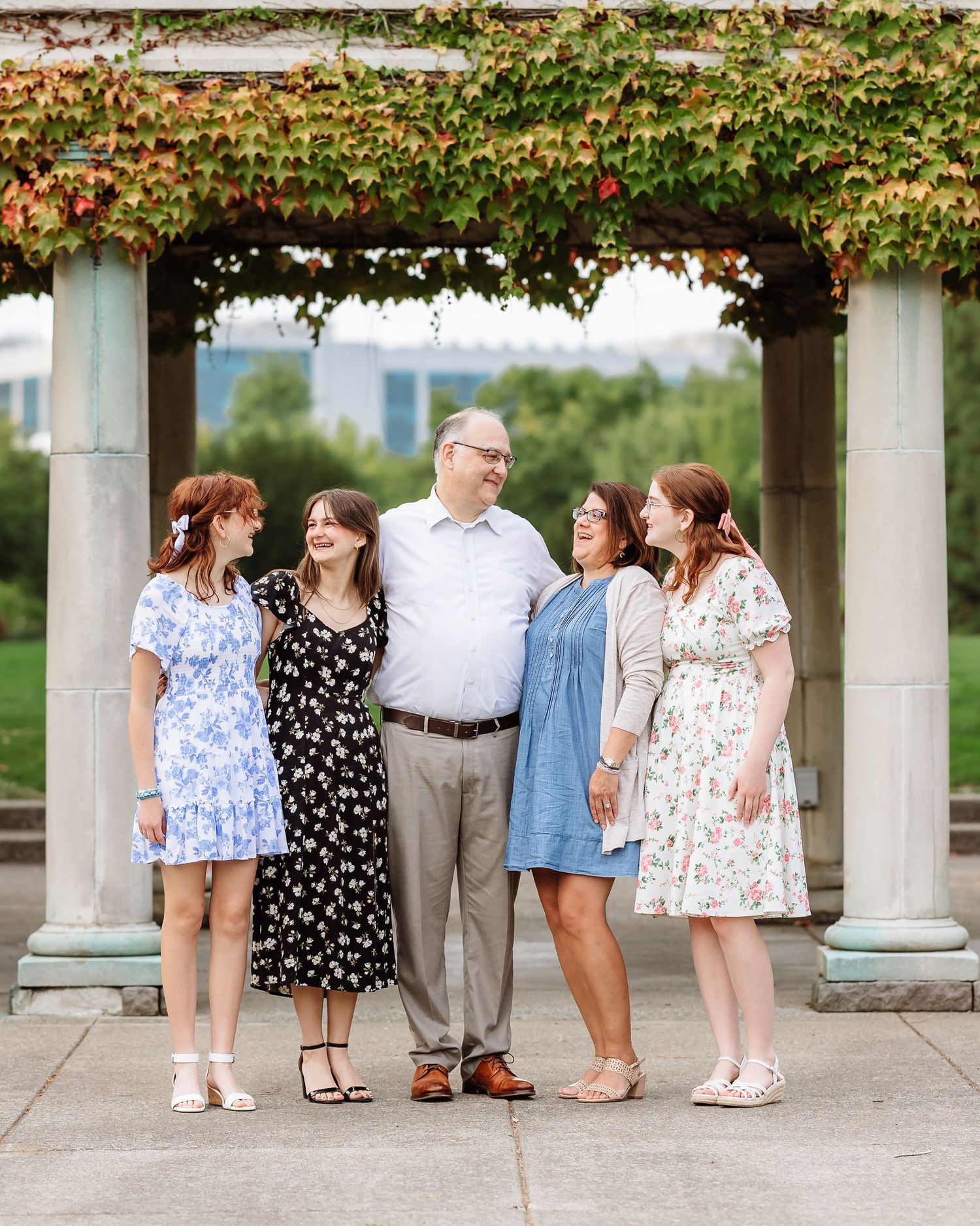 Family of five posing under an ivy-covered pergola in a bright summer outdoor portrait.