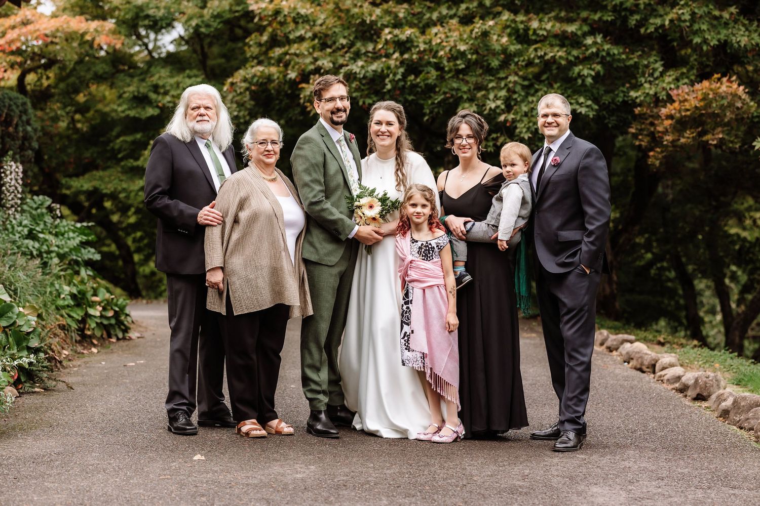 Wedding party family portrait outdoors on garden path surrounded by lush greenery.