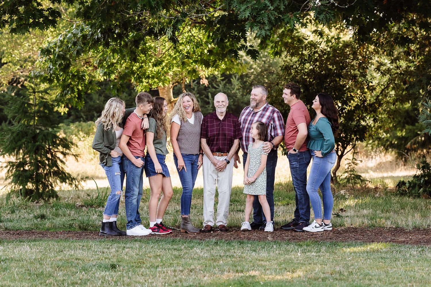 A family of seven smiling and laughing together outdoors on a sunny day surrounded by lush green trees.