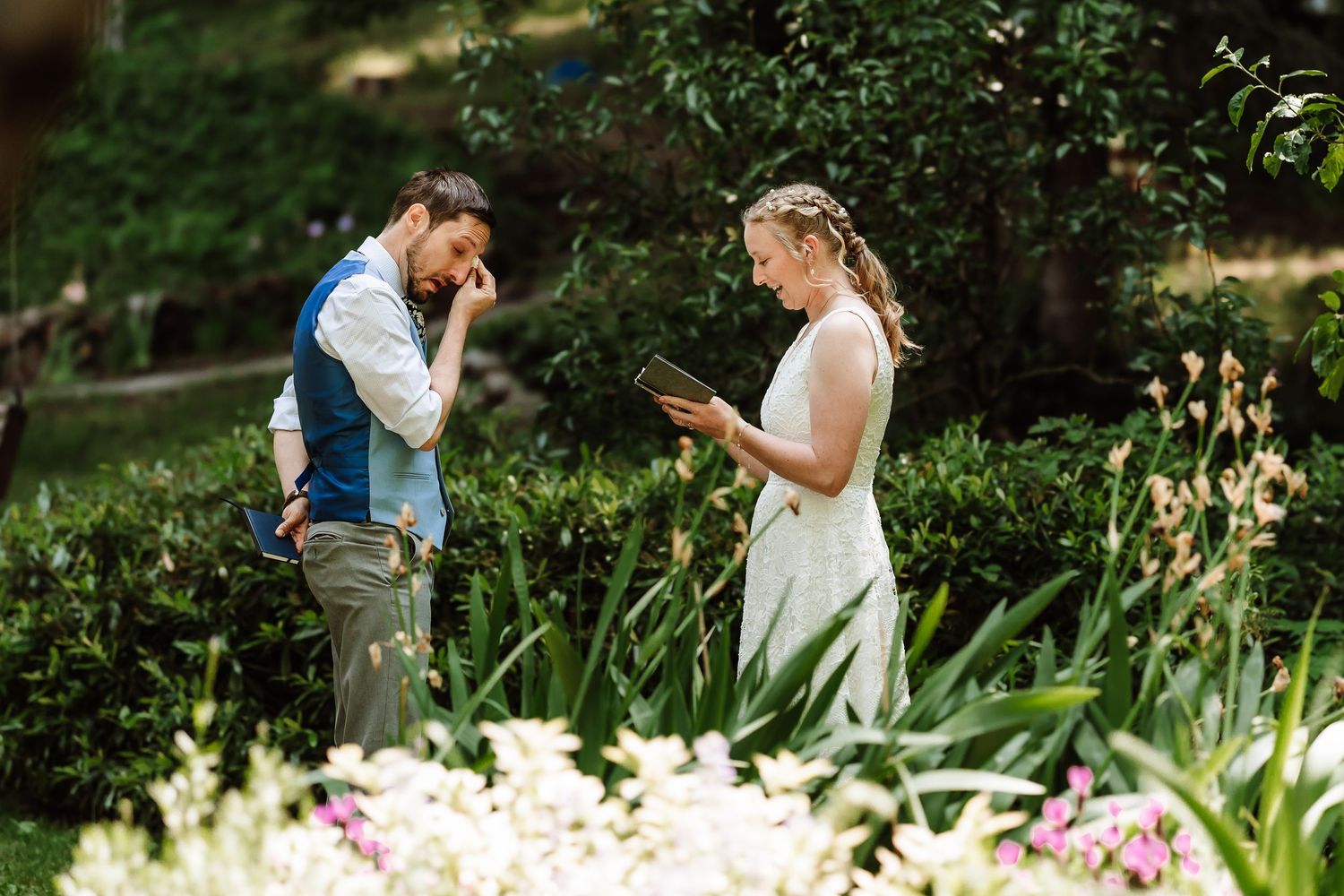 Emotional groom wiping tears as bride reads heartfelt vows in a lush garden ceremony.