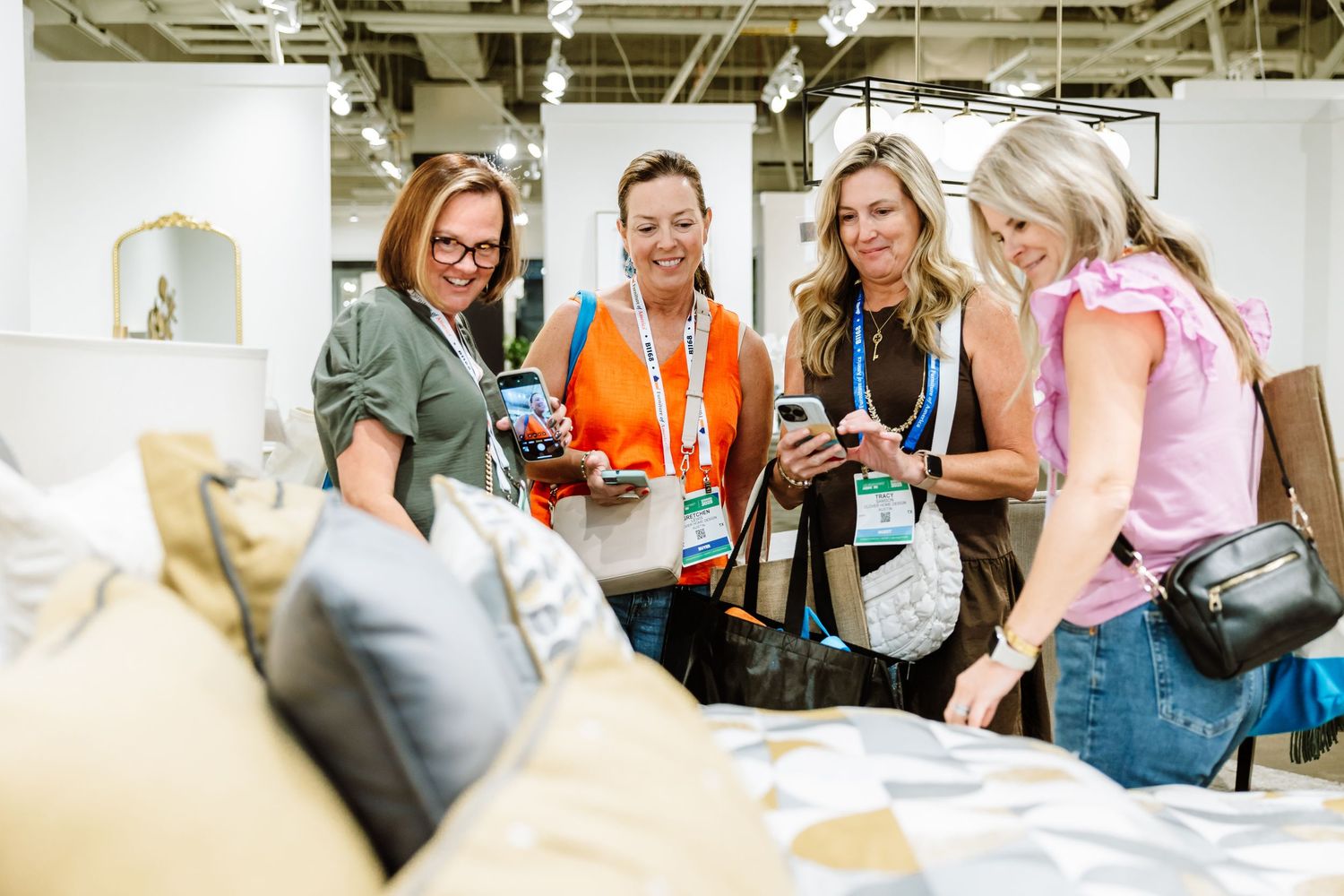 Four women smiling while browsing decorative pillows and home decor at interior design trade show.