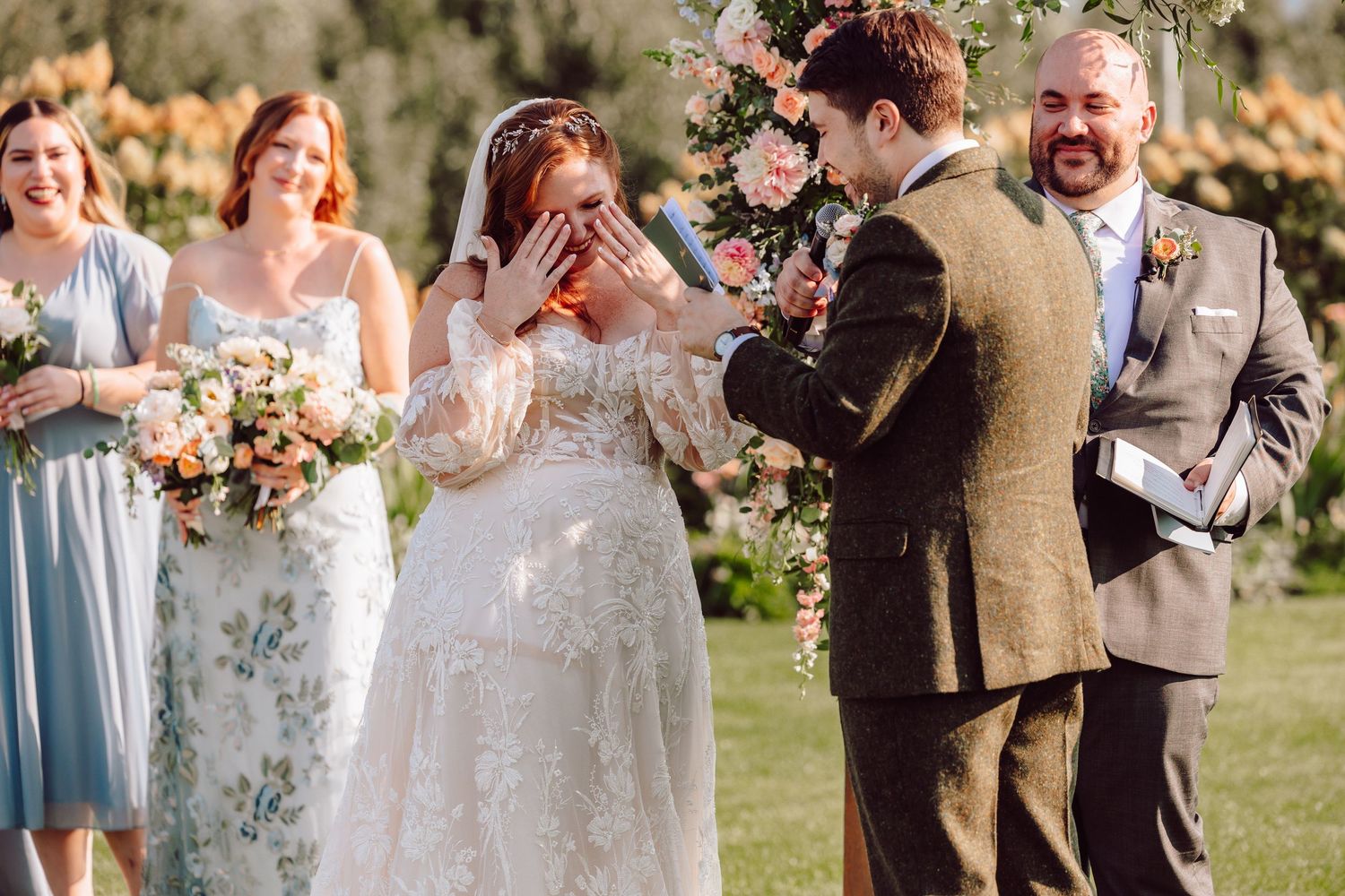 Emotional bride covers her face with joy as groom reads vows at outdoor wedding ceremony.