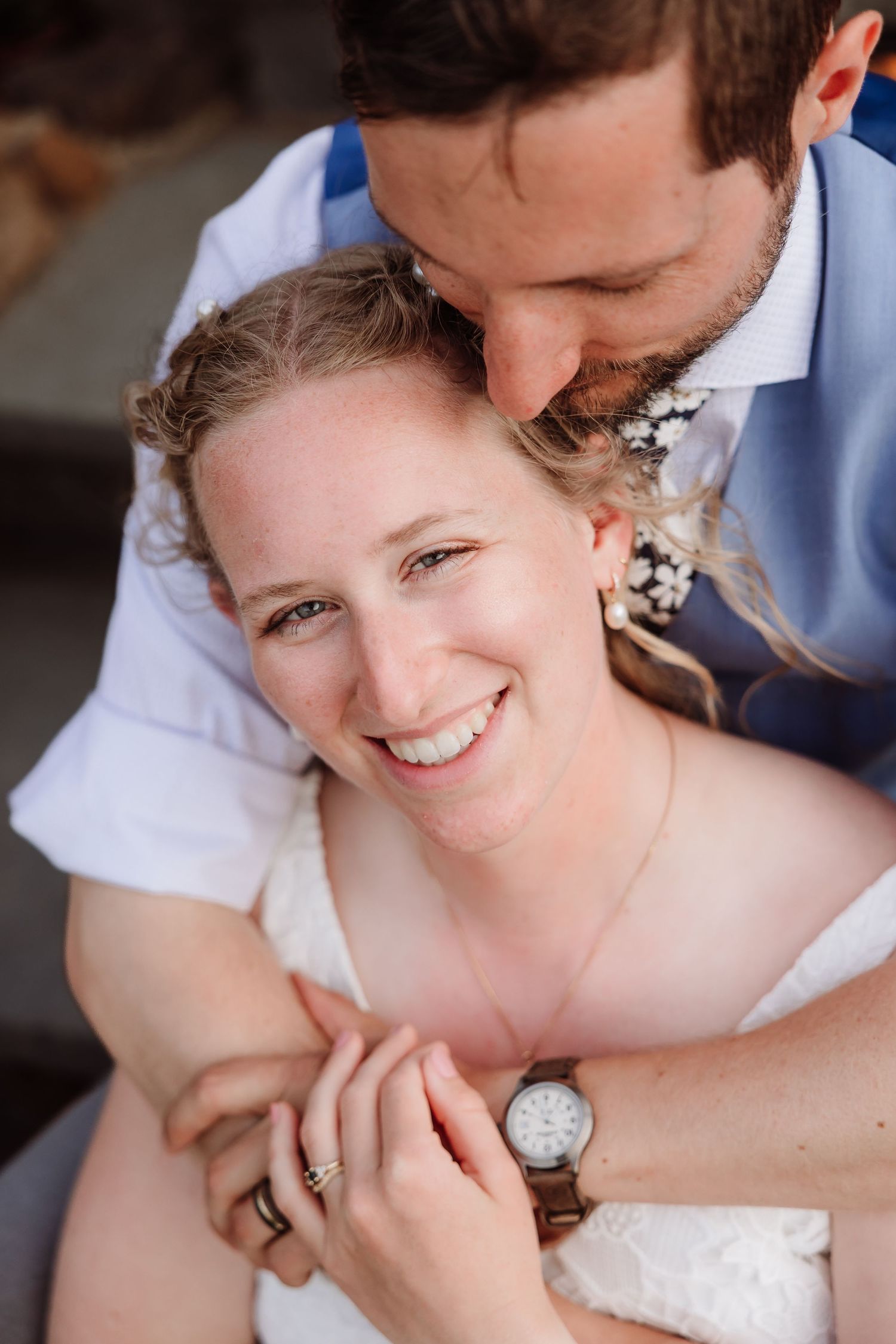 Groom embraces bride from behind as she smiles joyfully, wearing wedding attire outdoors.