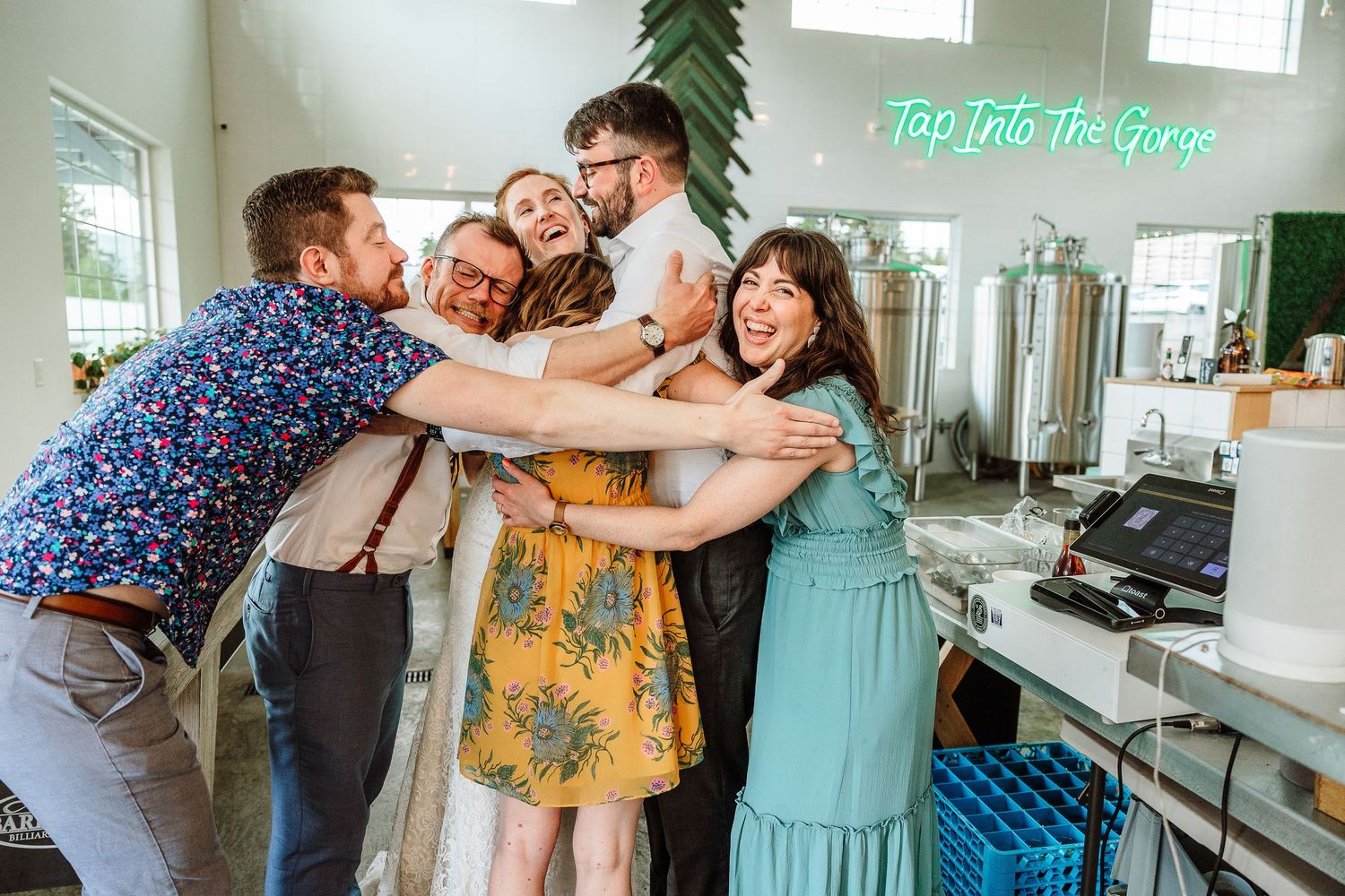 Bride and groom share group hug with wedding party friends inside brewery wedding reception venue.