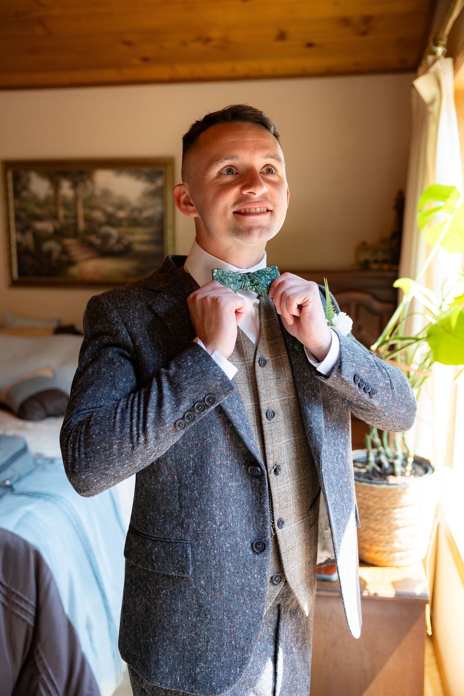 Smiling groom in a grey tweed suit adjusting his green floral bow tie before the wedding.