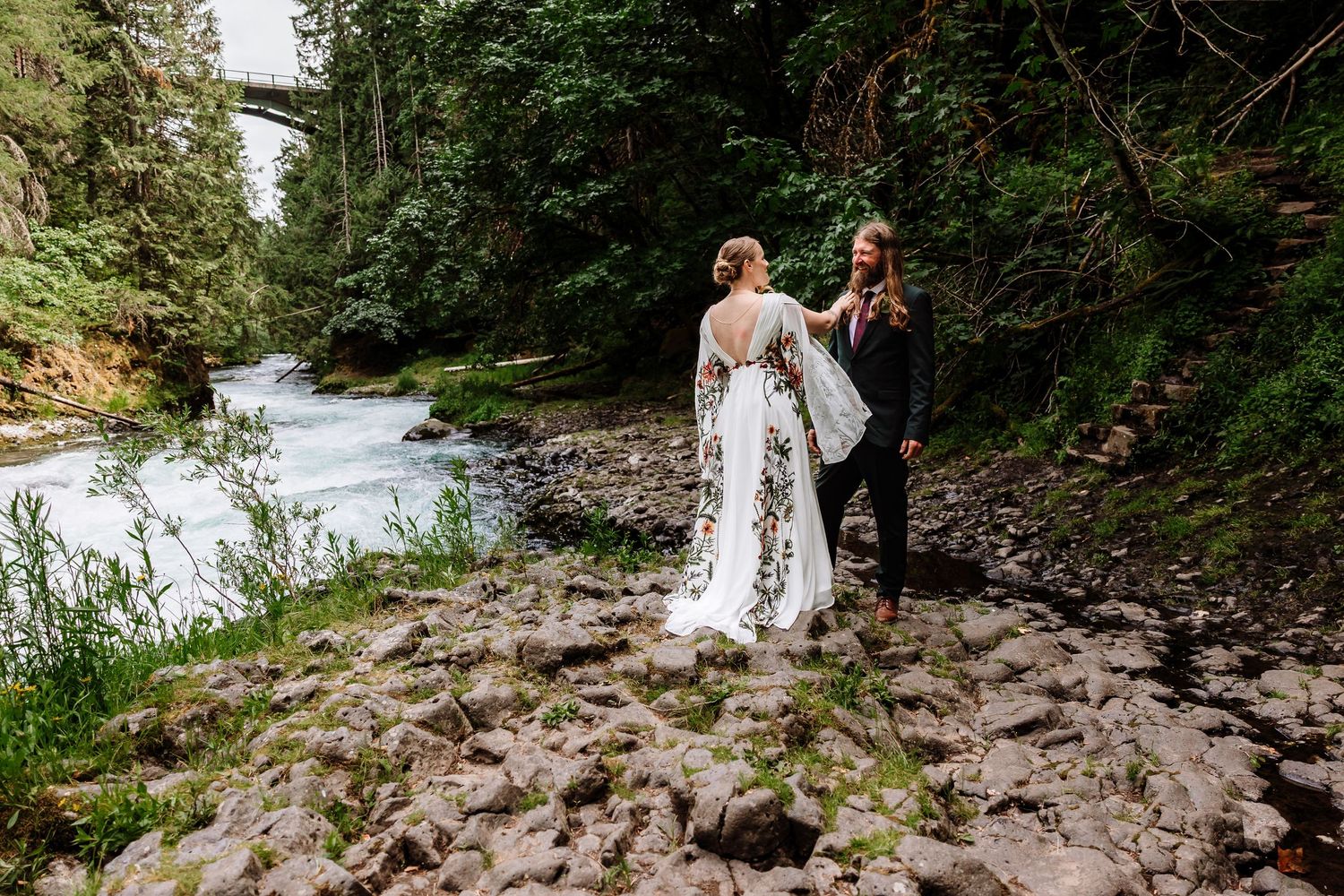 Bride and groom share an intimate moment on a rocky riverbank with a lush forest and river backdrop.