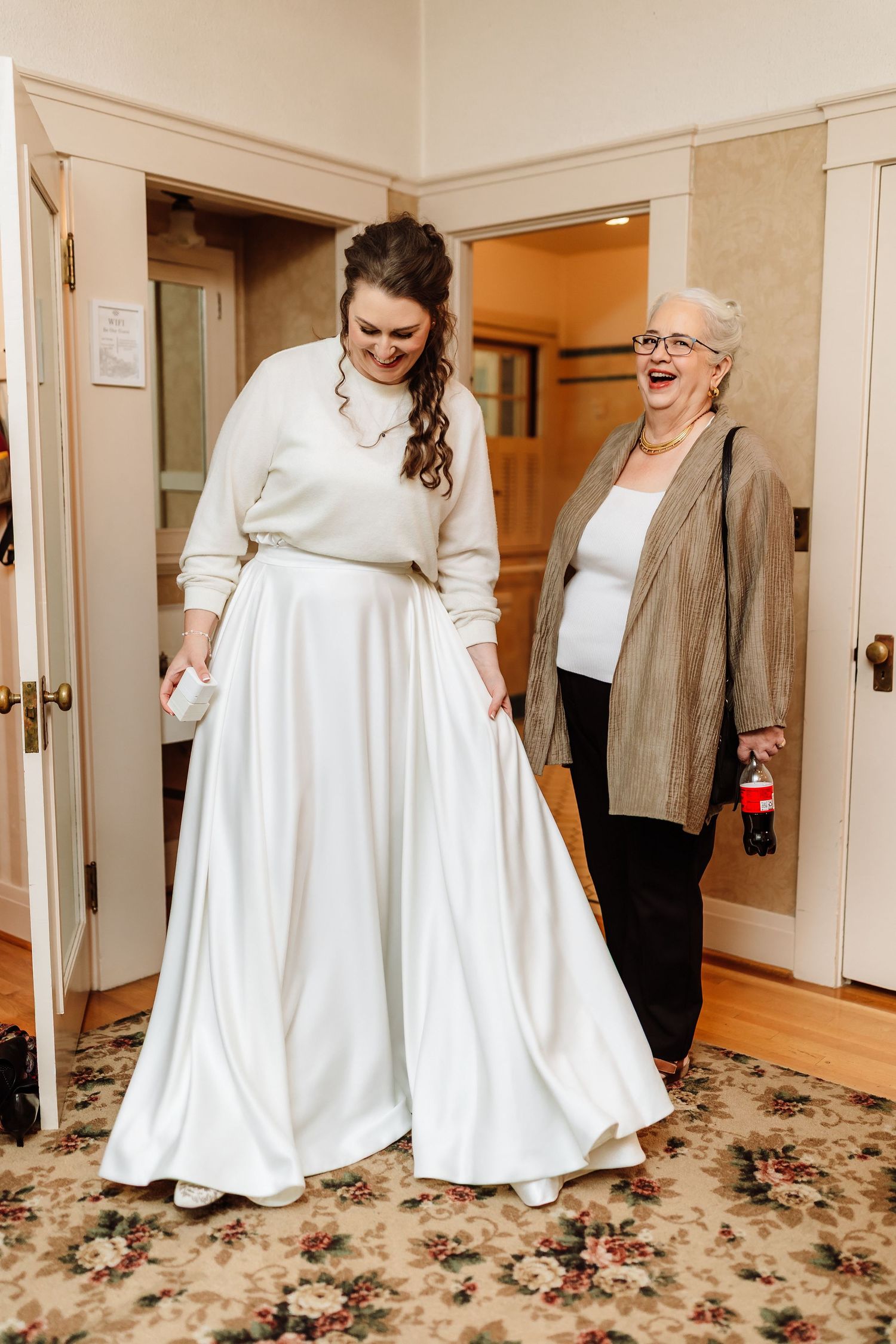 Bride in white satin skirt and sweater laughs with elderly woman in gold jacket during wedding preparations.