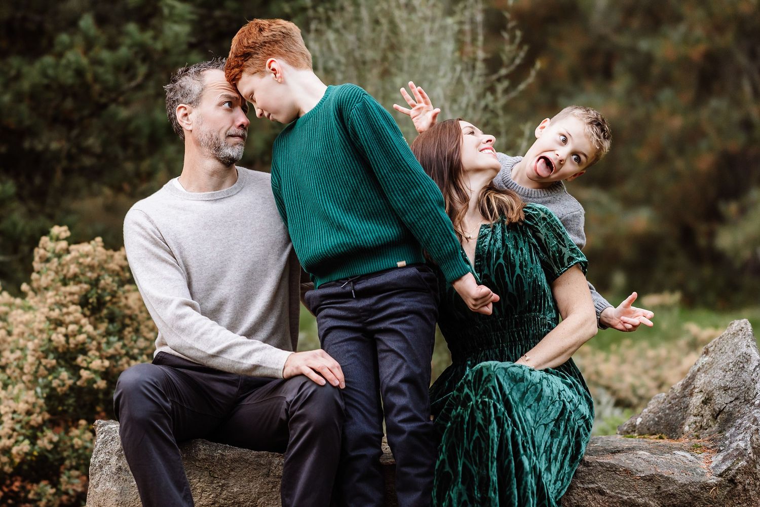 Playful family of four sitting on rocks outdoors, laughing together in a lush garden setting.