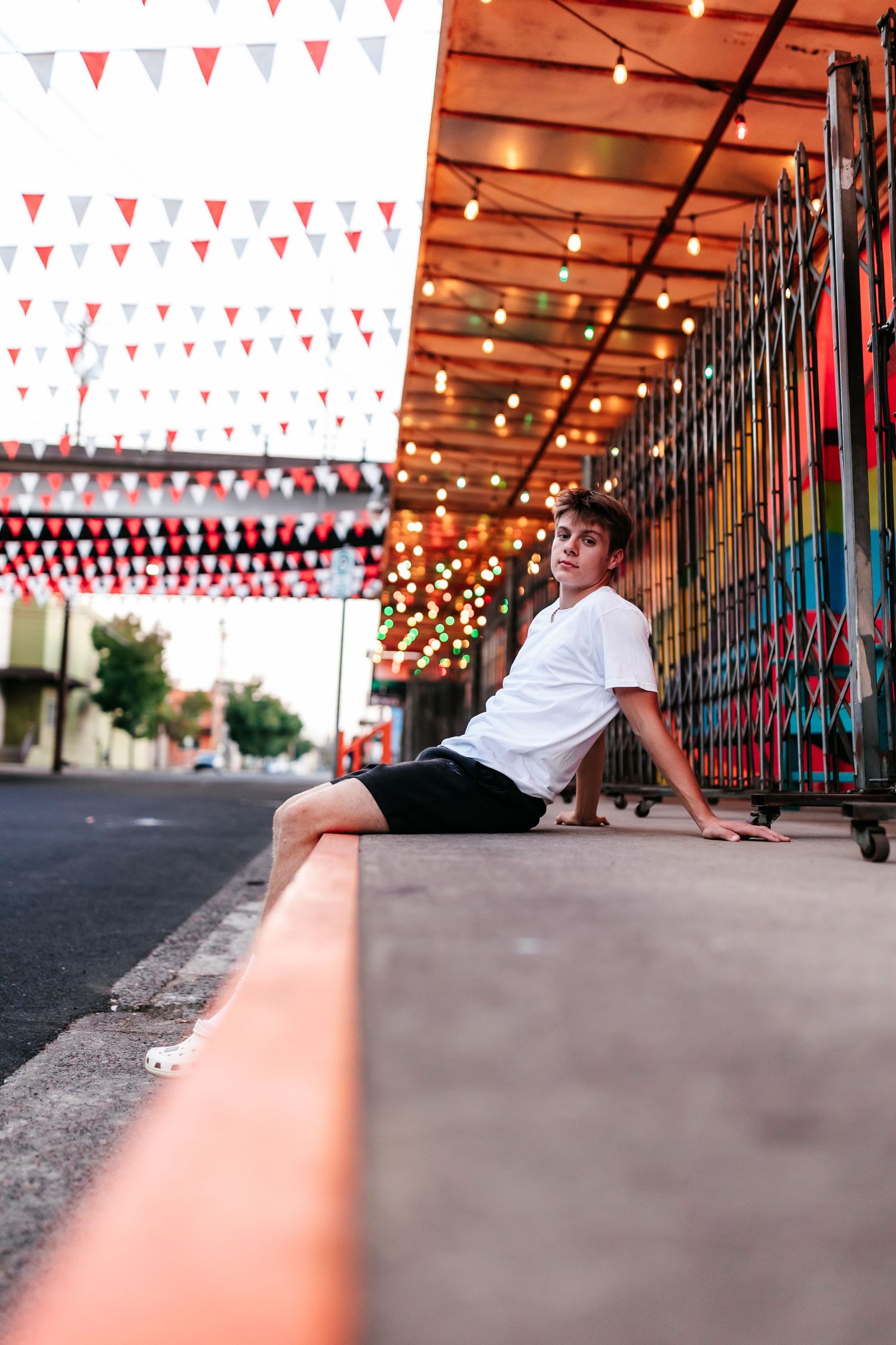 Young man in white t-shirt and black shorts sitting on a ledge under festive string lights and colorful bunting.