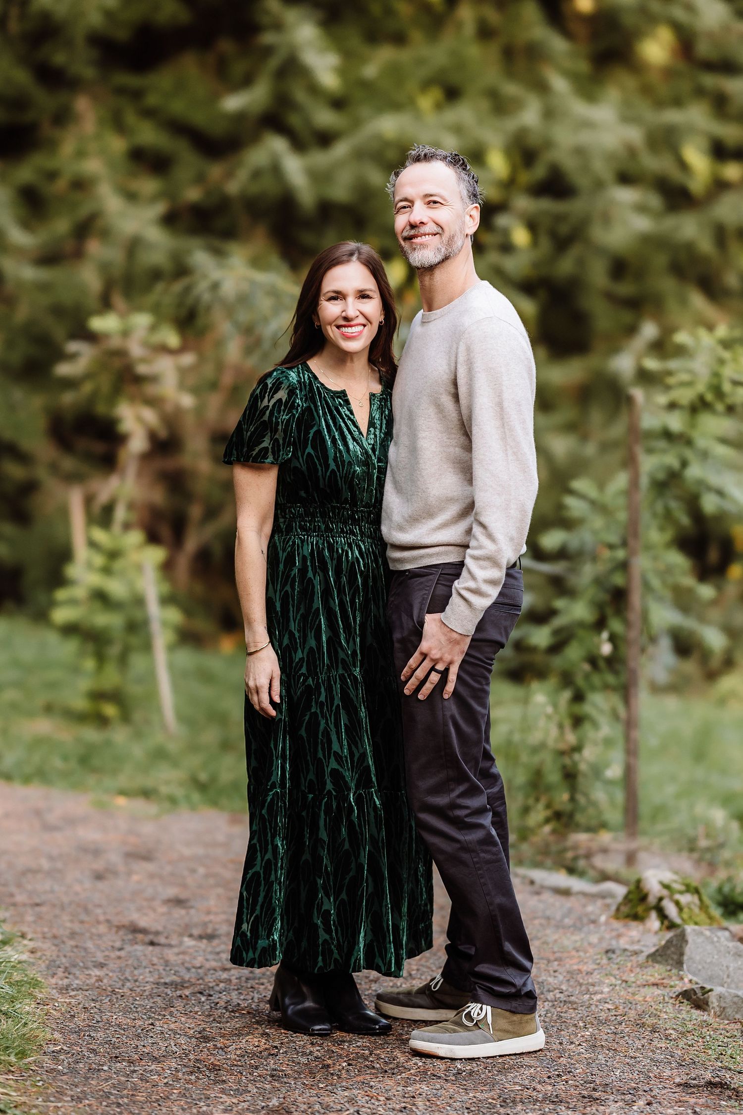 Smiling couple posing outdoors on a garden path, woman in green velvet dress and man in beige sweater with dark pants.