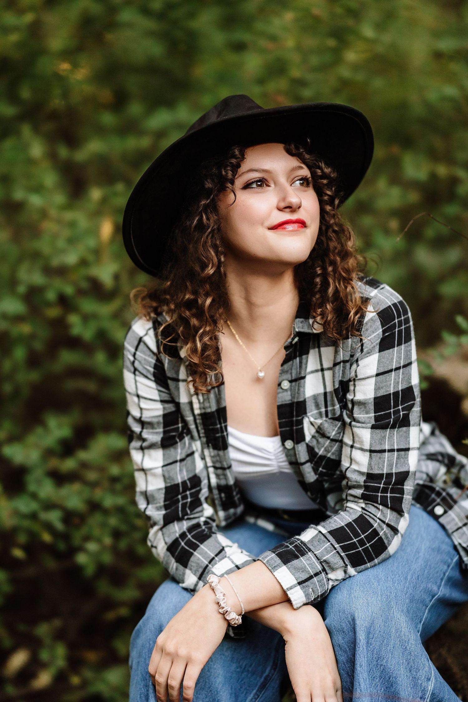 Young woman with curly hair wearing a black hat and plaid shirt sits outdoors, smiling thoughtfully.