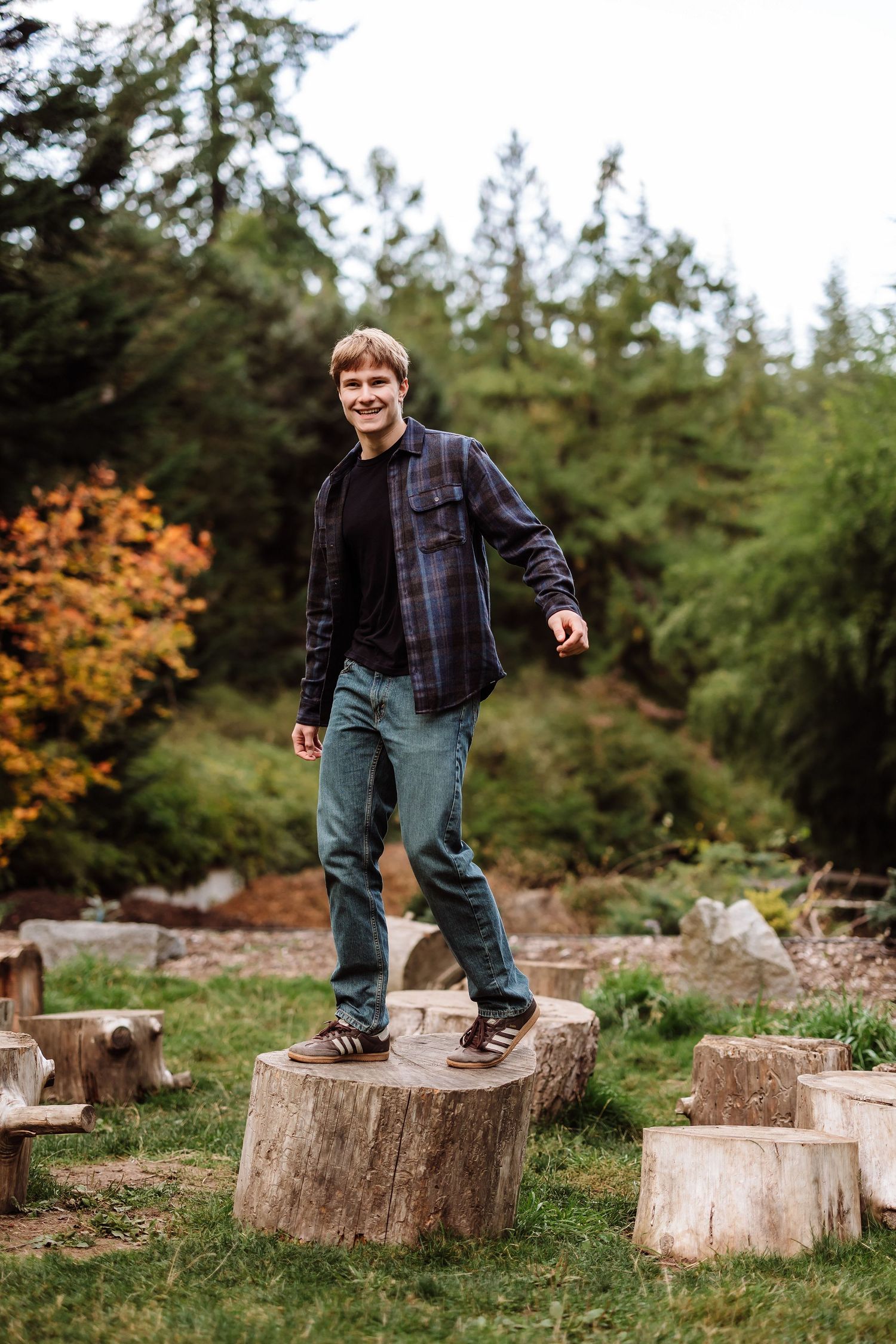Young man smiling while balancing on tree stumps in an outdoor park setting surrounded by autumn trees.