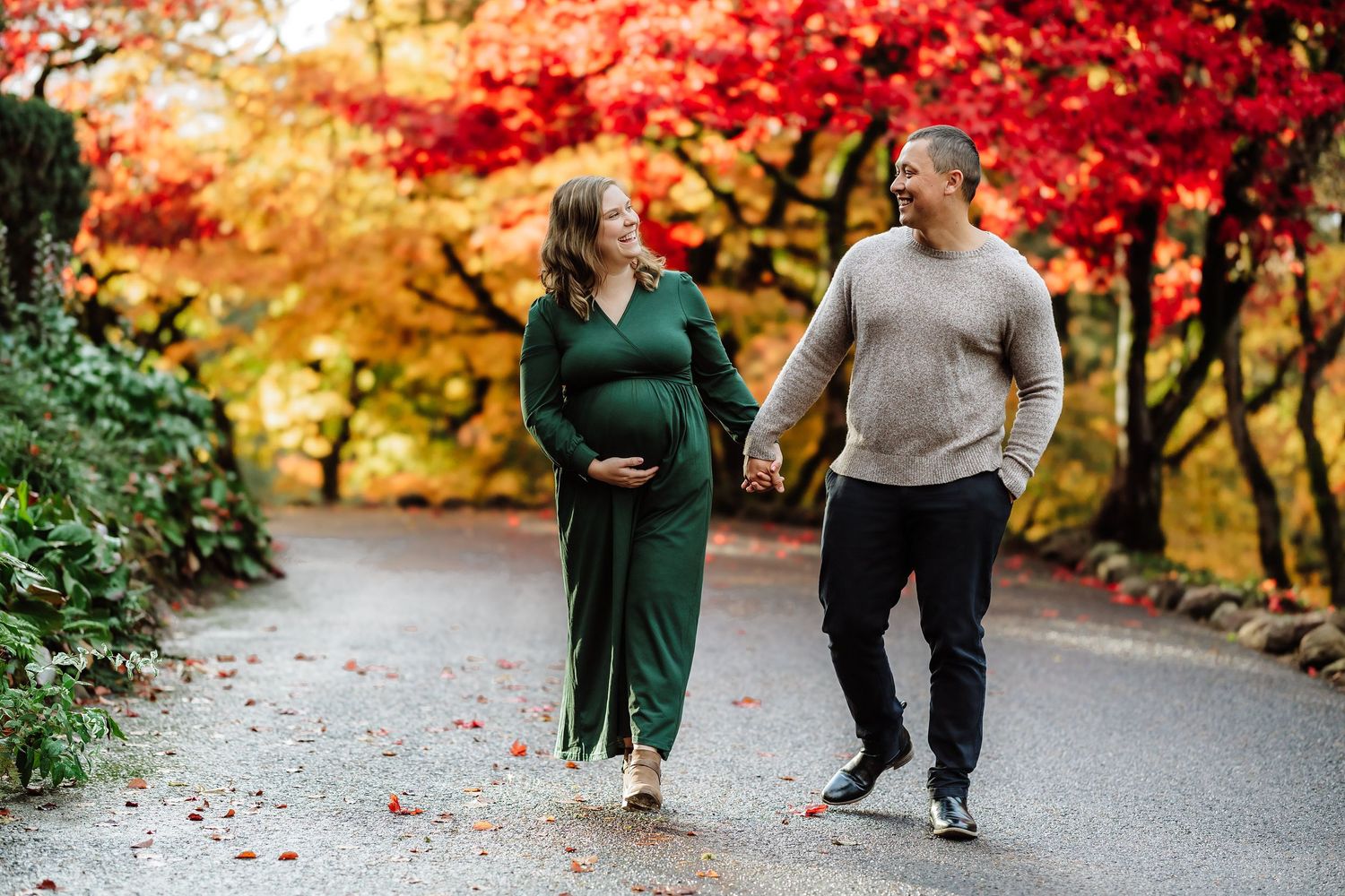 Pregnant couple holding hands on a tree-lined path surrounded by vibrant red and orange autumn foliage.