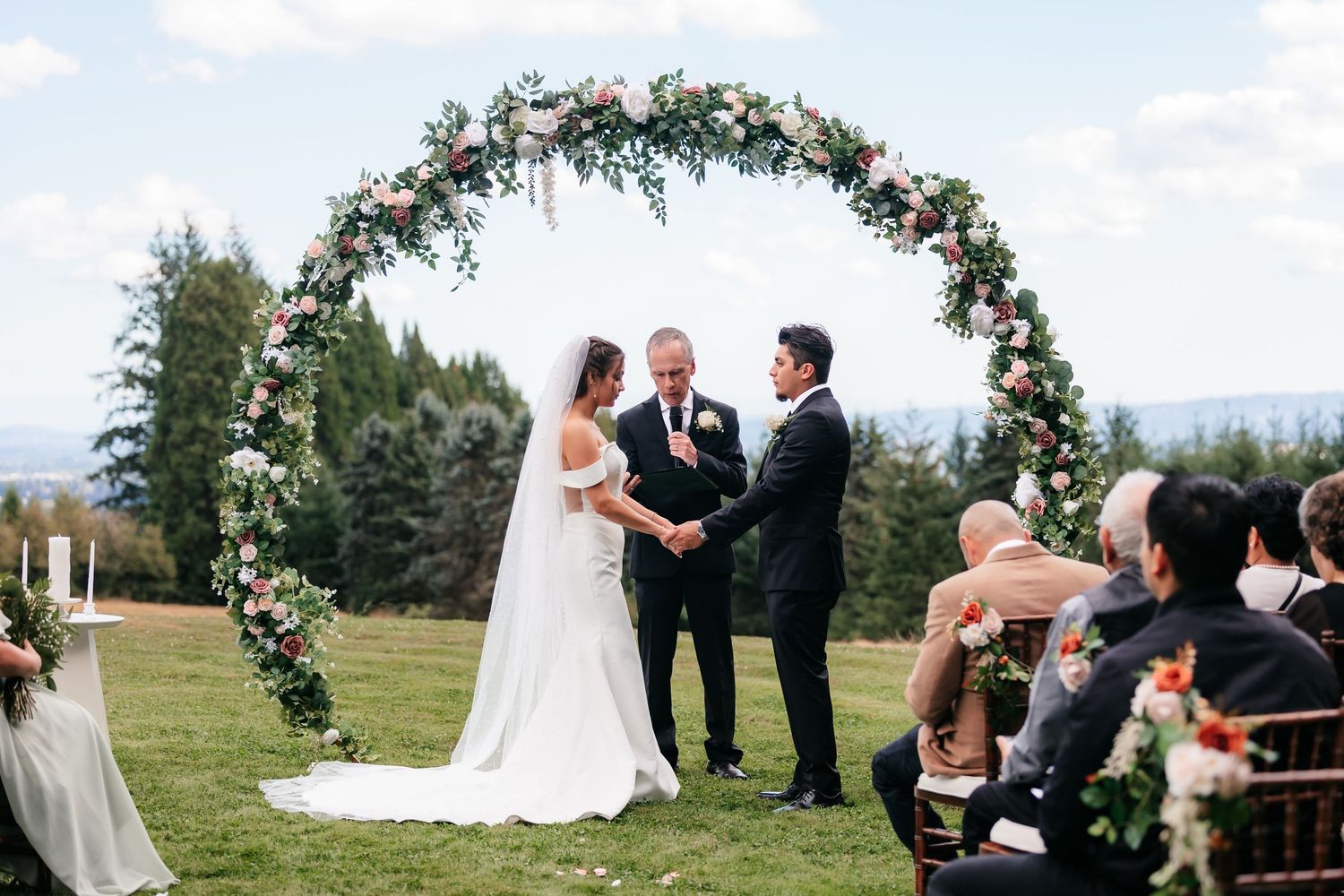 Bride and groom exchange vows under a lush circular floral arch at a scenic outdoor hilltop wedding.