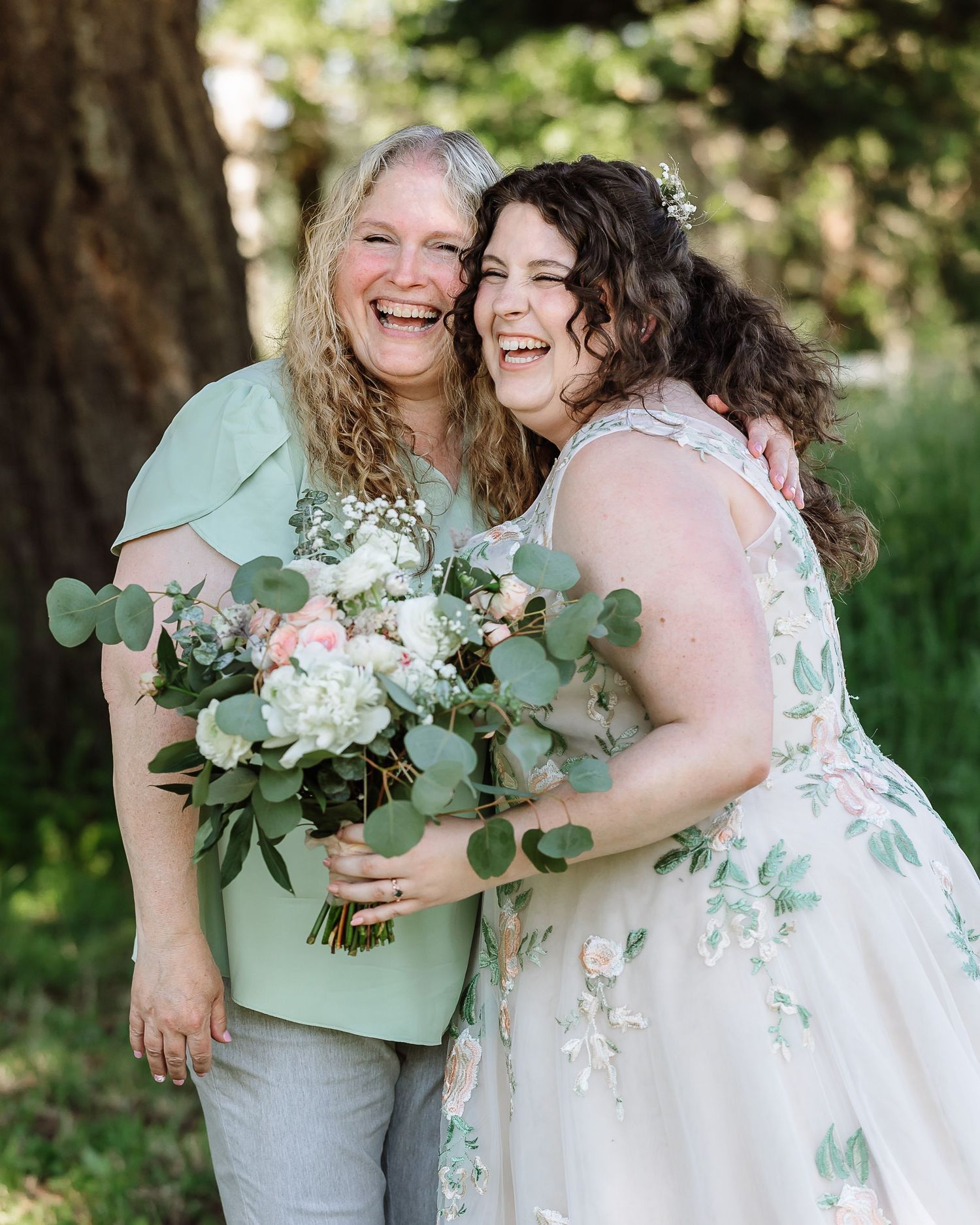 Bride in floral gown laughs joyfully with woman in mint green top, holding white and pink bouquet outdoors.